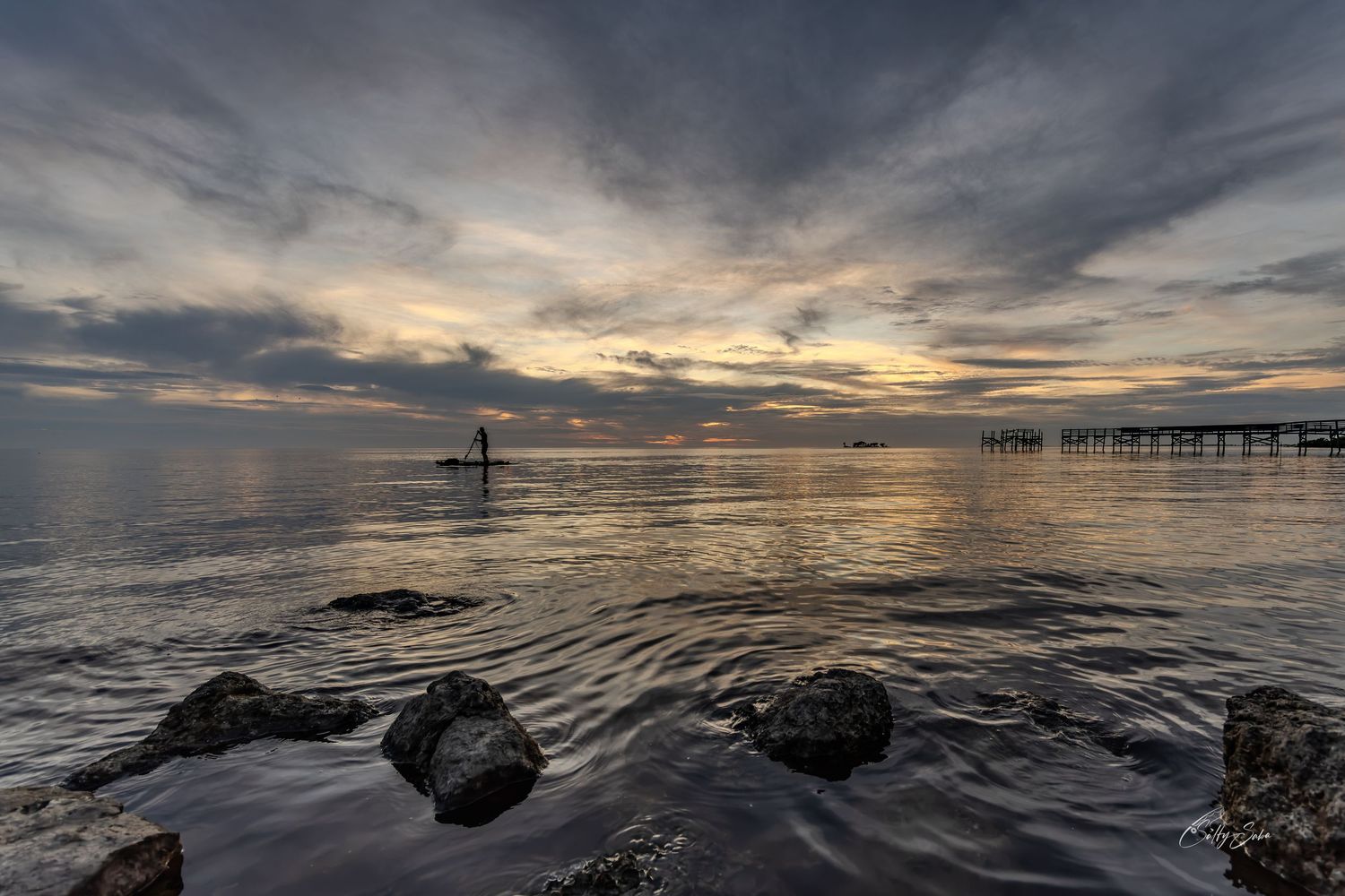 A silhouetted paddleboarder at sunset, with shoreline rocks in the foreground and a pier to the right.