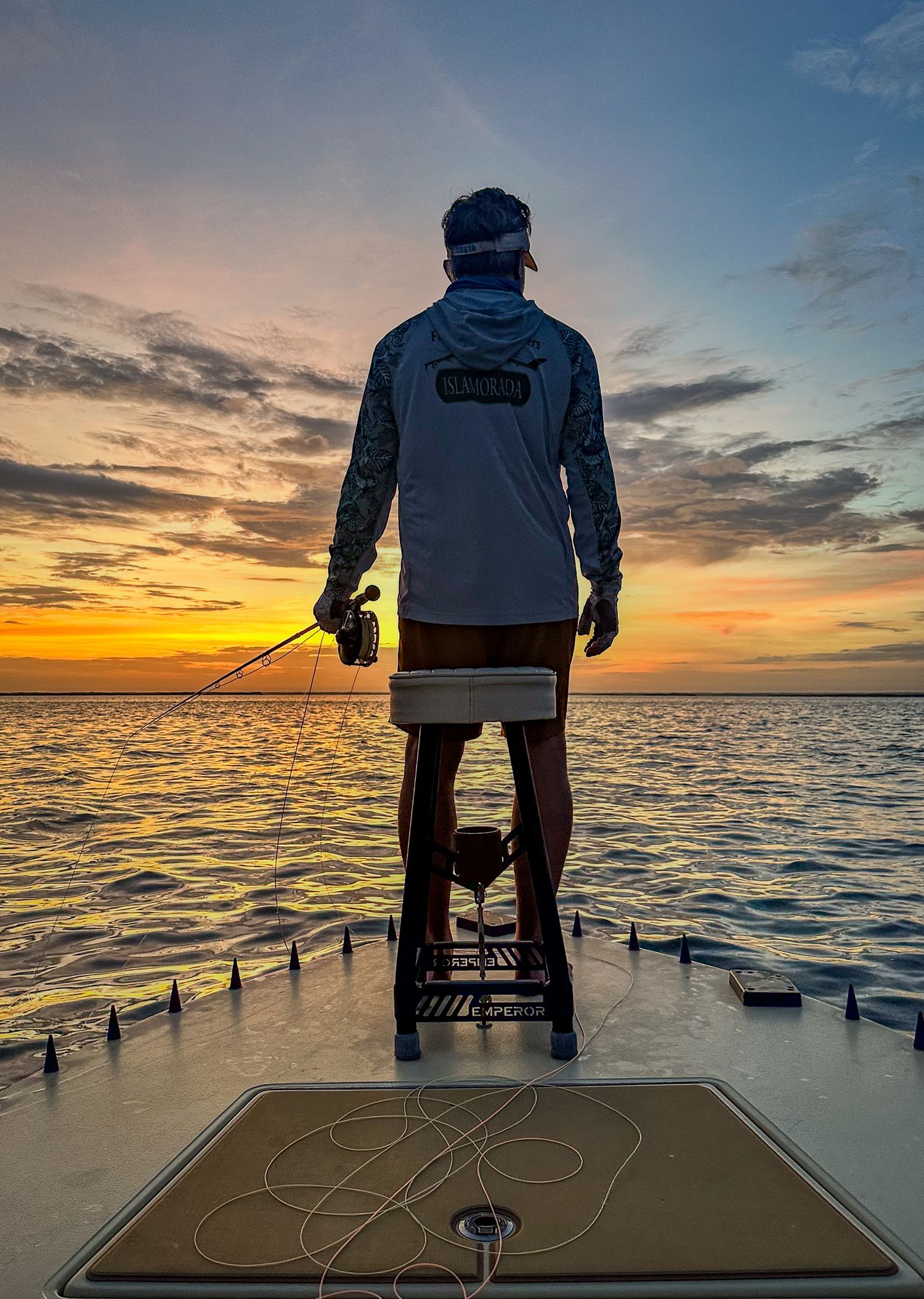 A fly-fishing angler stands on a skiff at sunrise with rod and line ready.