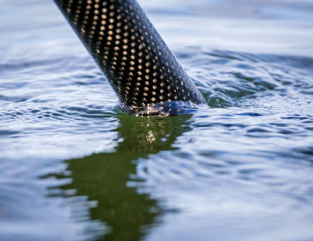 A dark carbon-fiber pole enters rippling blue water, its reflection distorted across the surface.