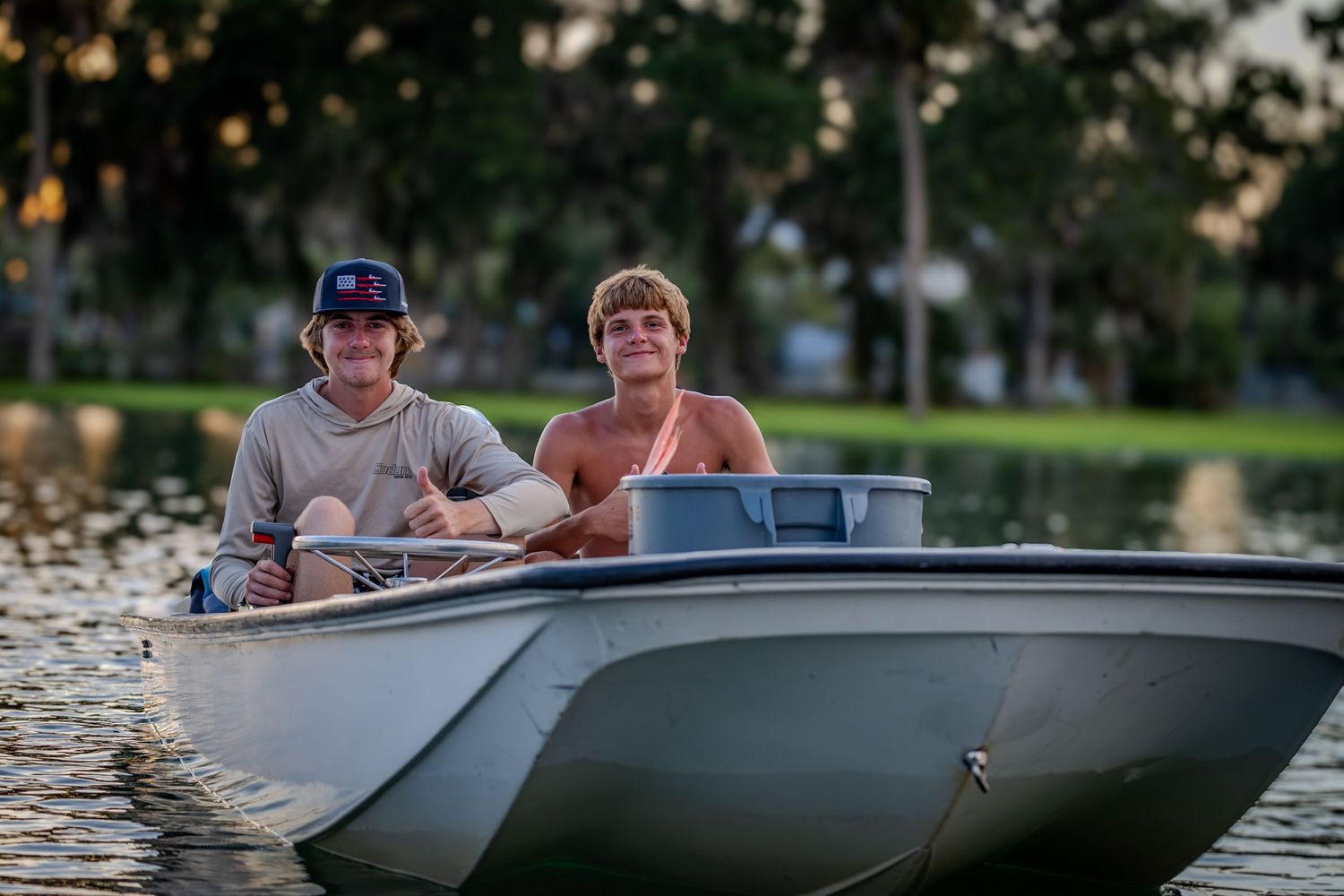 Two young men smile from a small skiff on calm water in evening light.