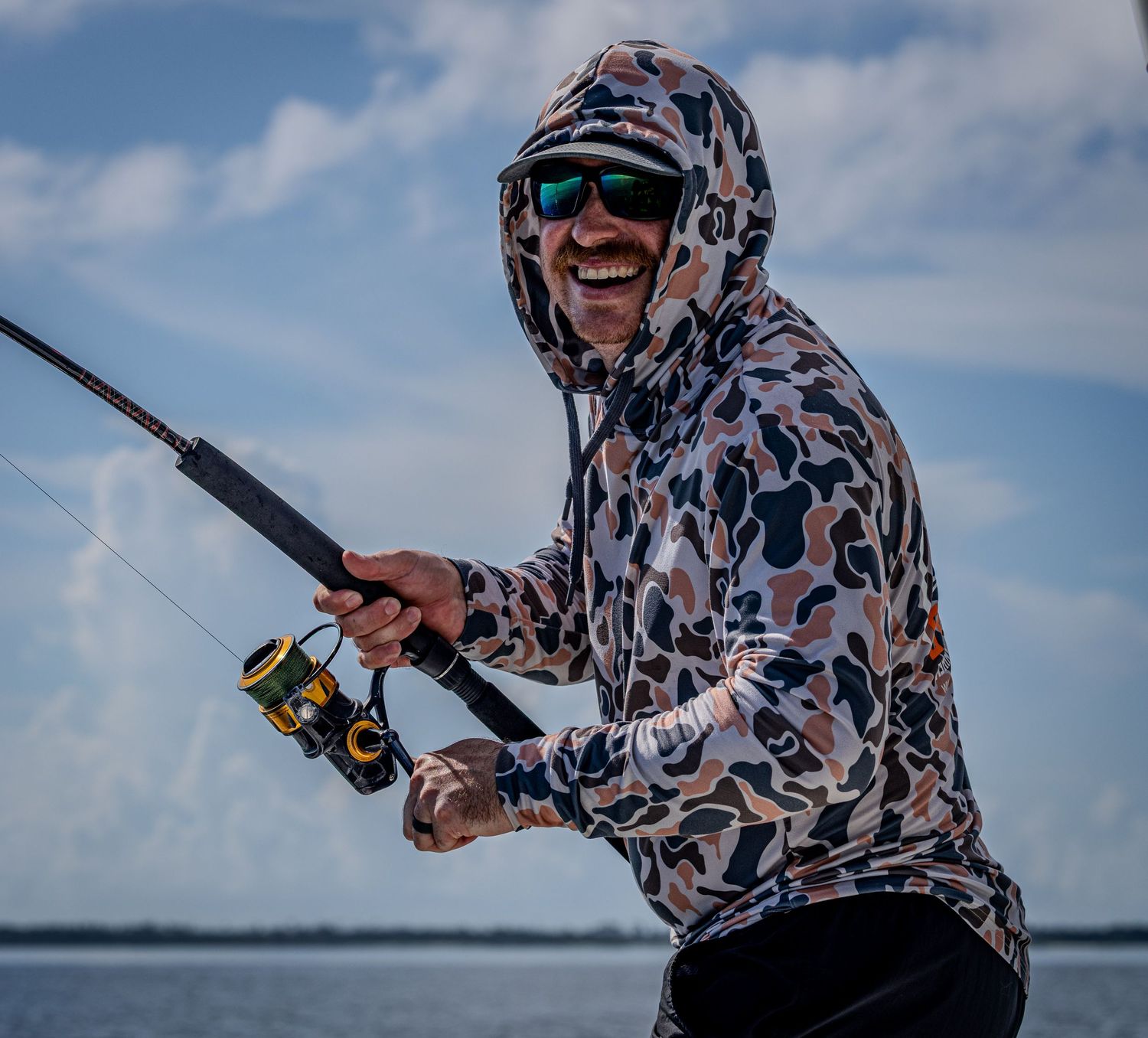 A smiling angler in a camouflage sun hoodie holds a spinning rod beneath a blue sky.
