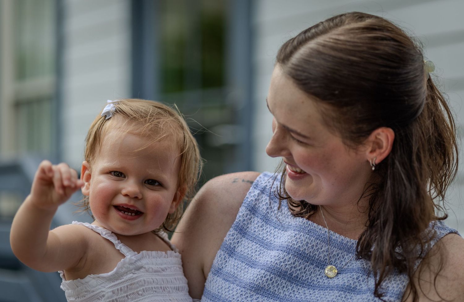 A woman holds a smiling toddler outdoors as they look toward one another.