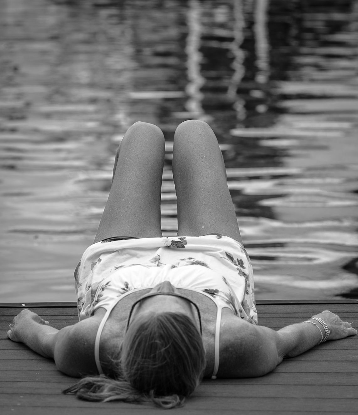 Black-and-white photograph of a woman lying back on a dock beside rippling water.