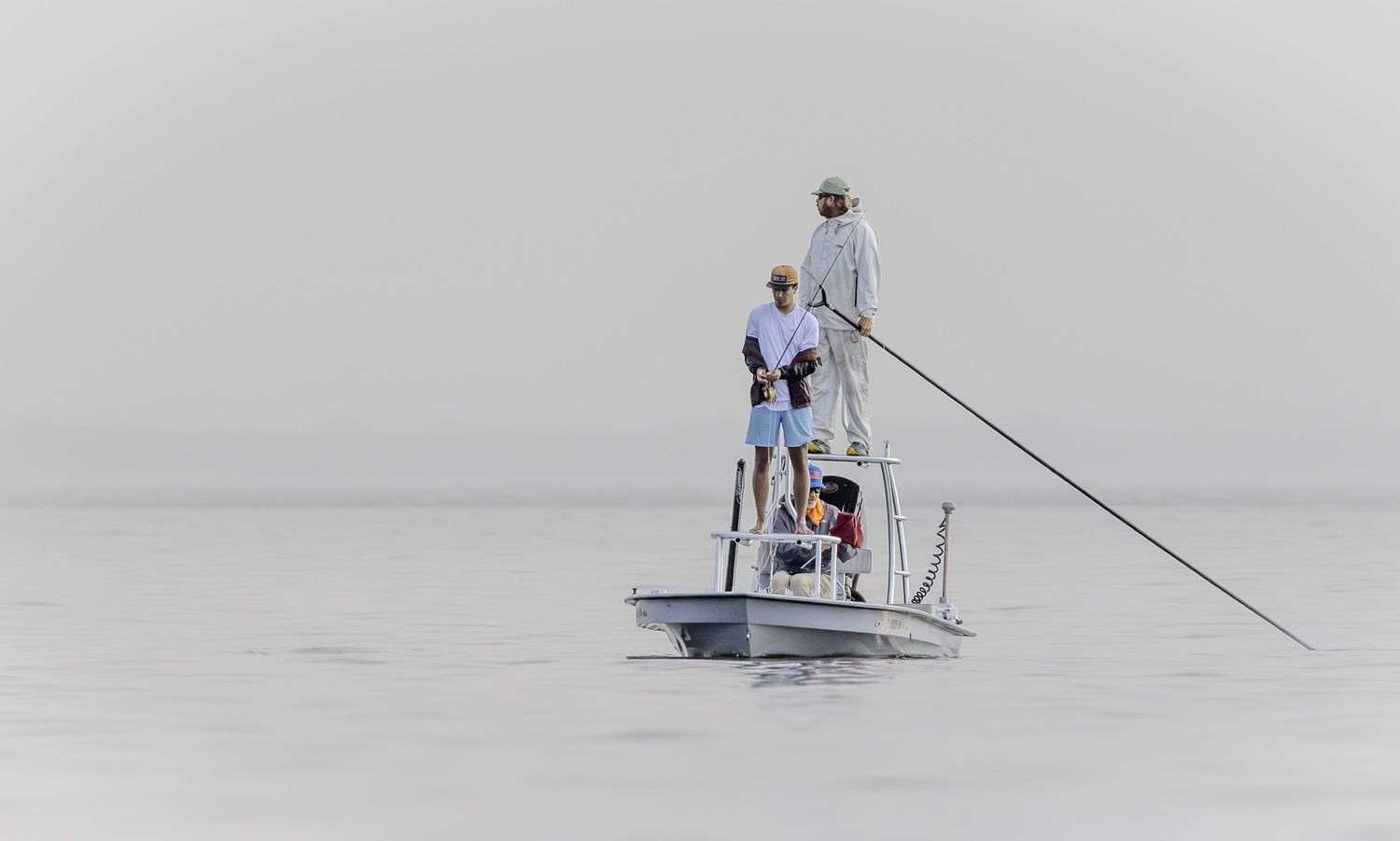 A guide poles a flats skiff carrying young anglers through pale morning haze.