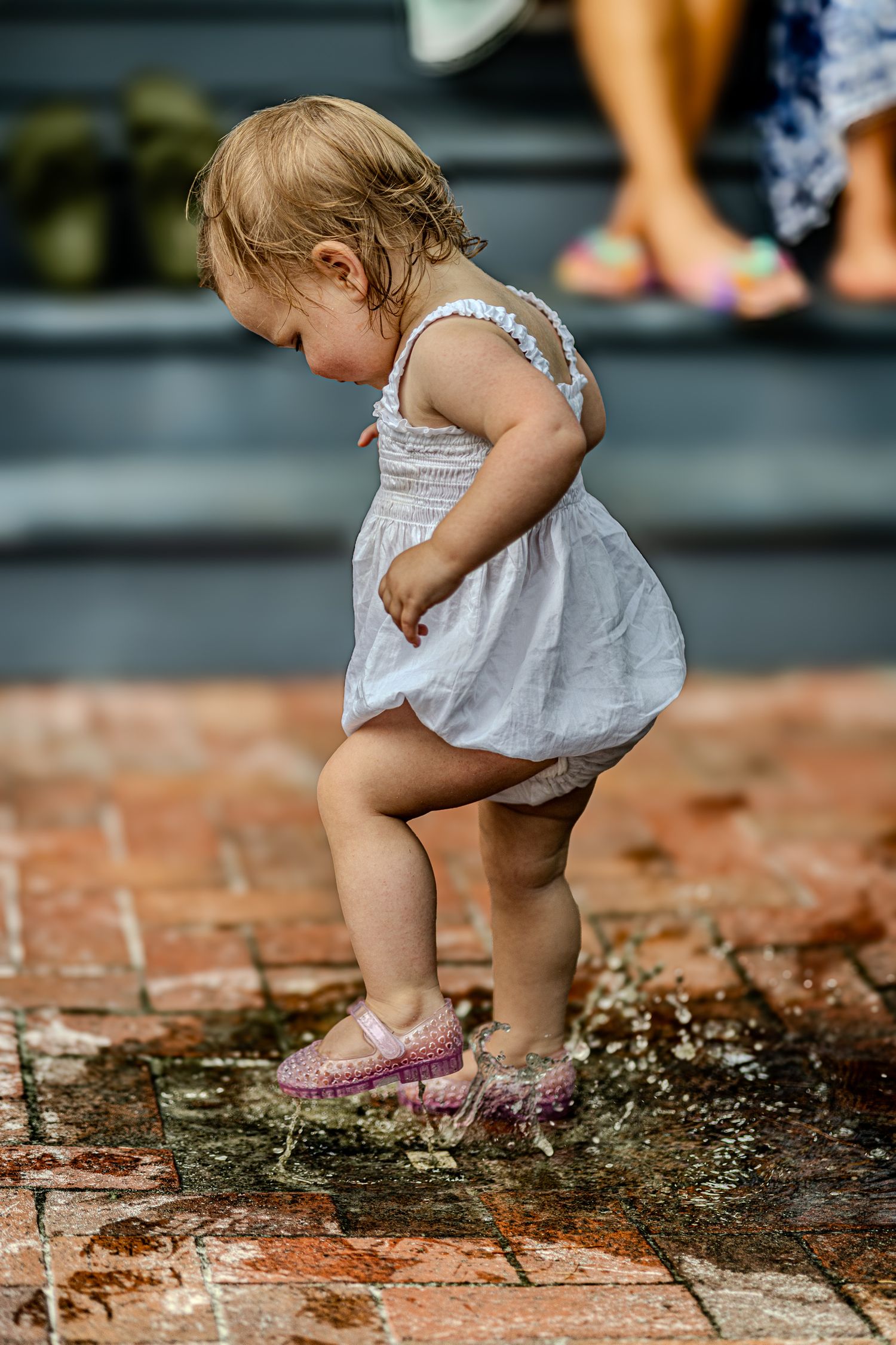A toddler in a white dress splashes through a shallow puddle on a brick patio.