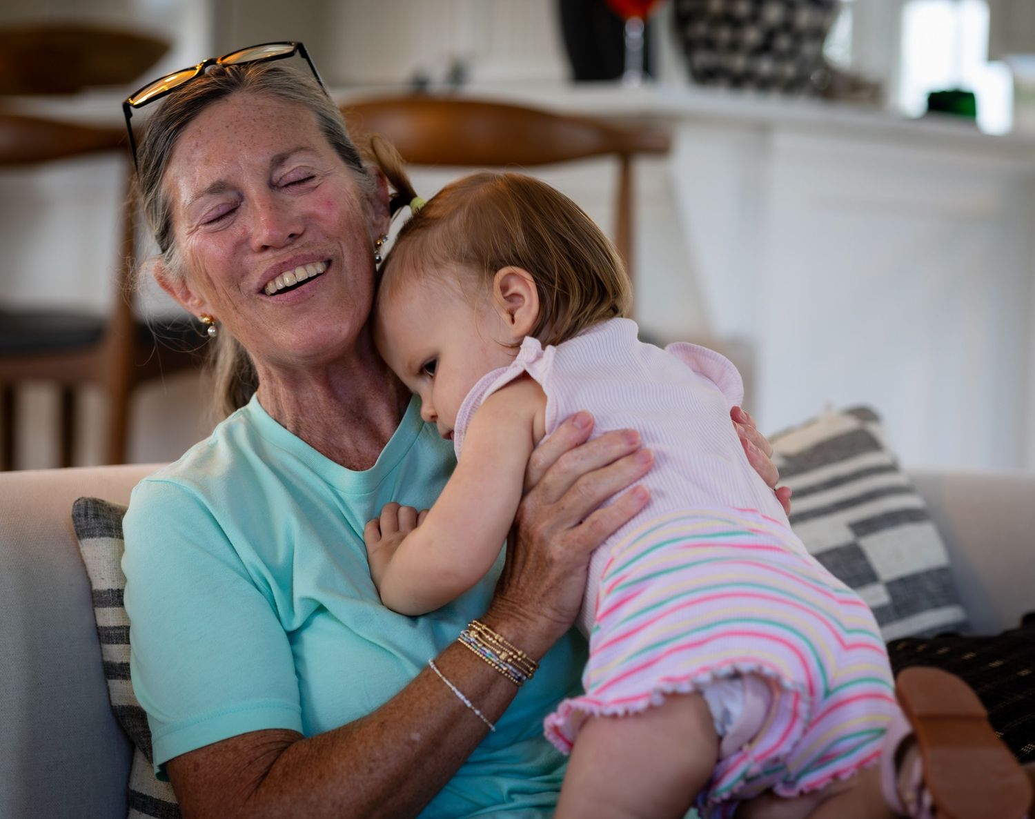 A woman closes her eyes and smiles while embracing a baby on a sofa.