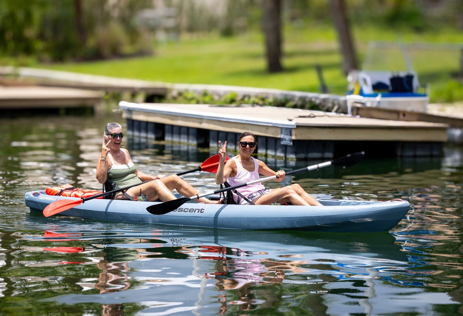 Two women in a tandem kayak smile and gesture while paddling on calm water.