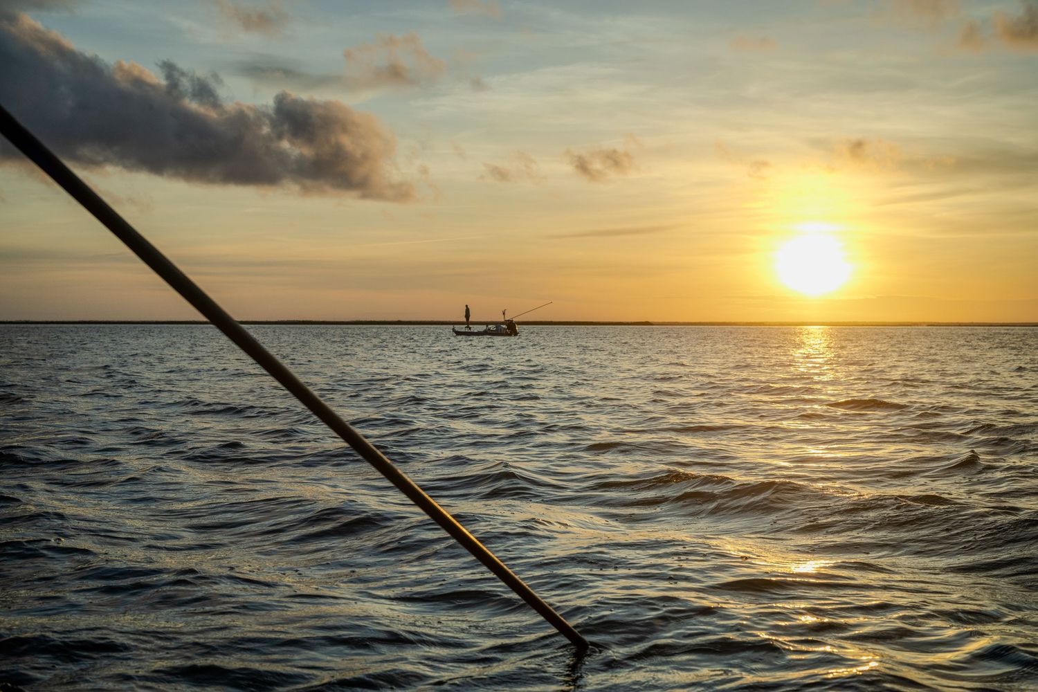 A distant flats skiff at sunrise, framed by a diagonal push pole over rippling water.