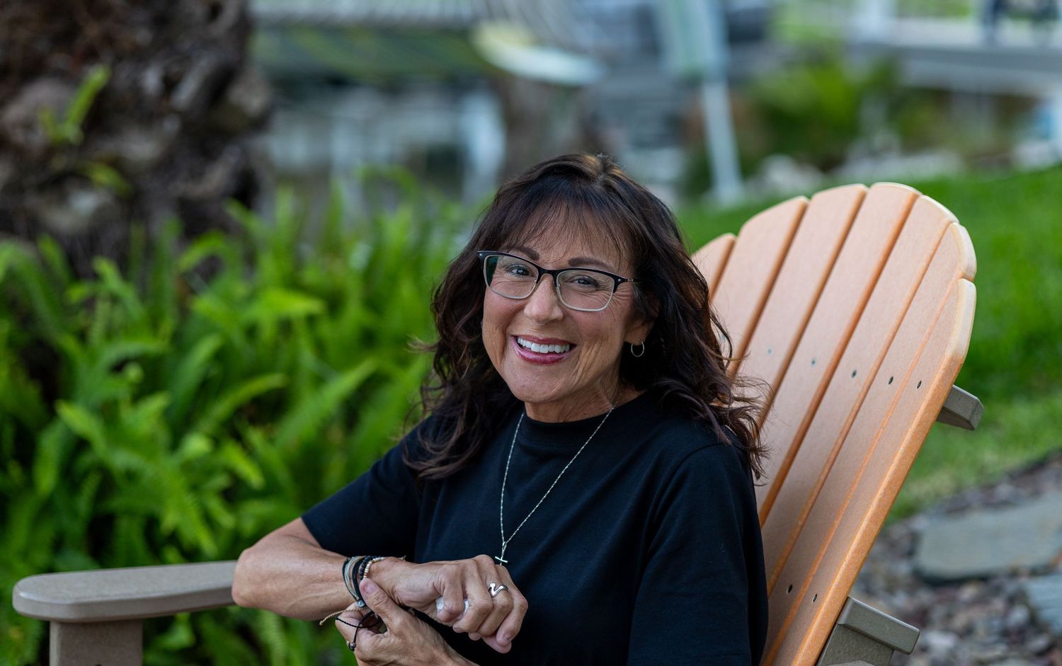 A smiling woman in glasses sits in an orange Adirondack chair beside lush green ferns.