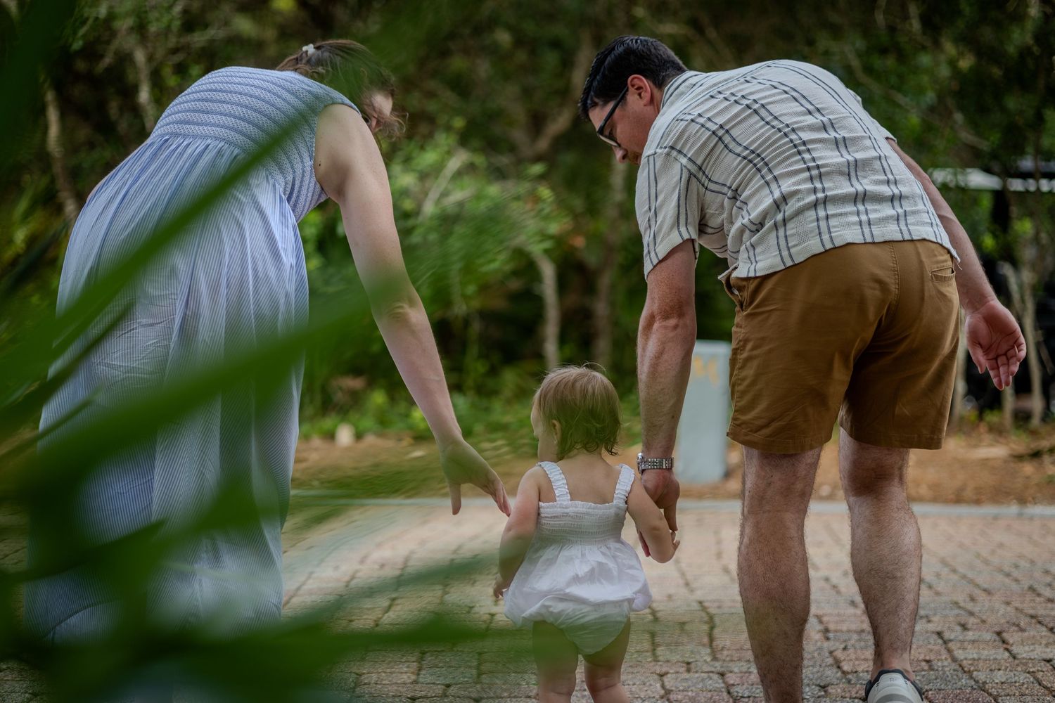 Two adults guide a toddler along a shaded brick path, seen from behind.