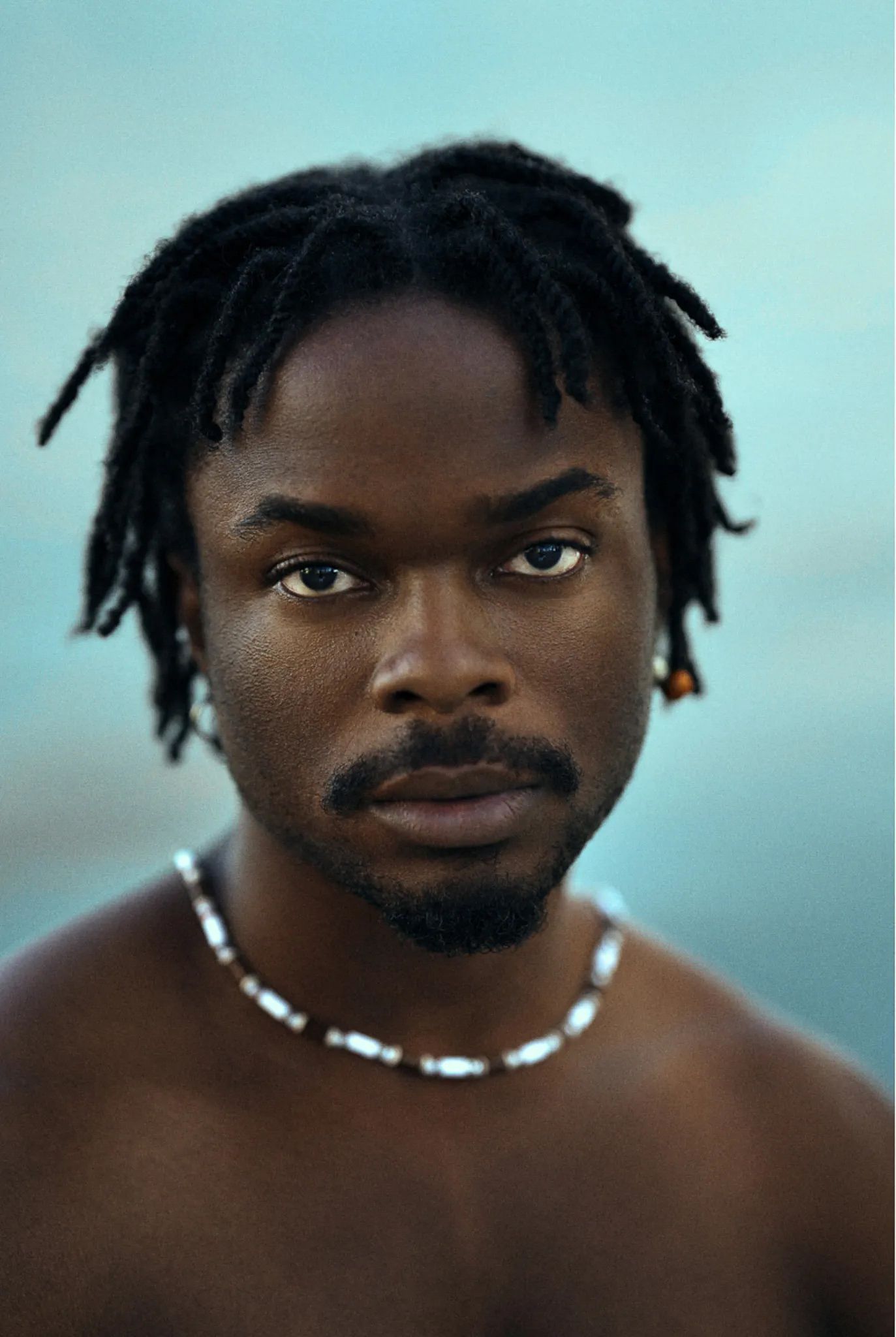 Young Black man with dreadlocks and beaded necklace against a soft blue background.