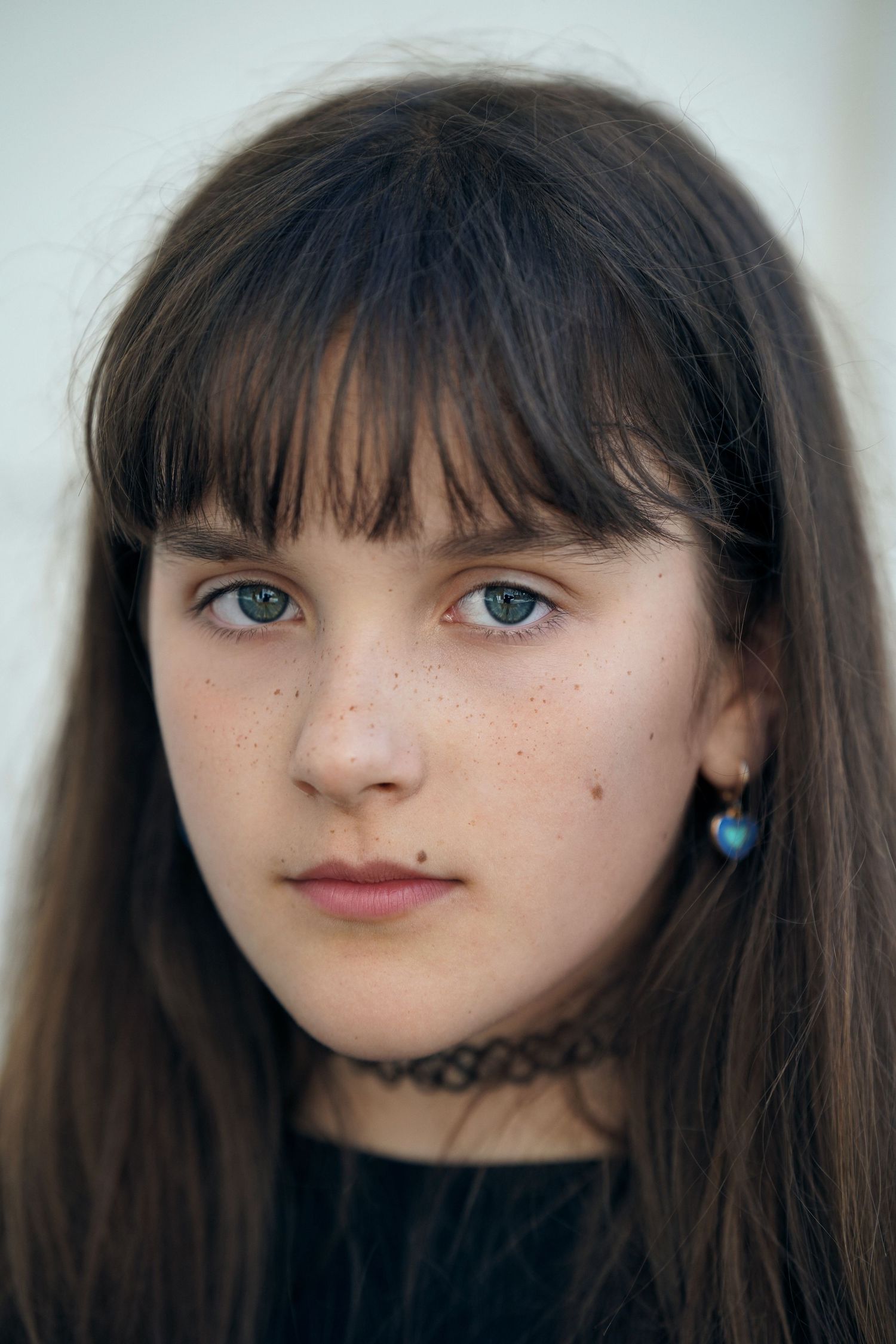Close-up portrait of a young girl with dark hair, bangs, freckles, and blue heart earrings.