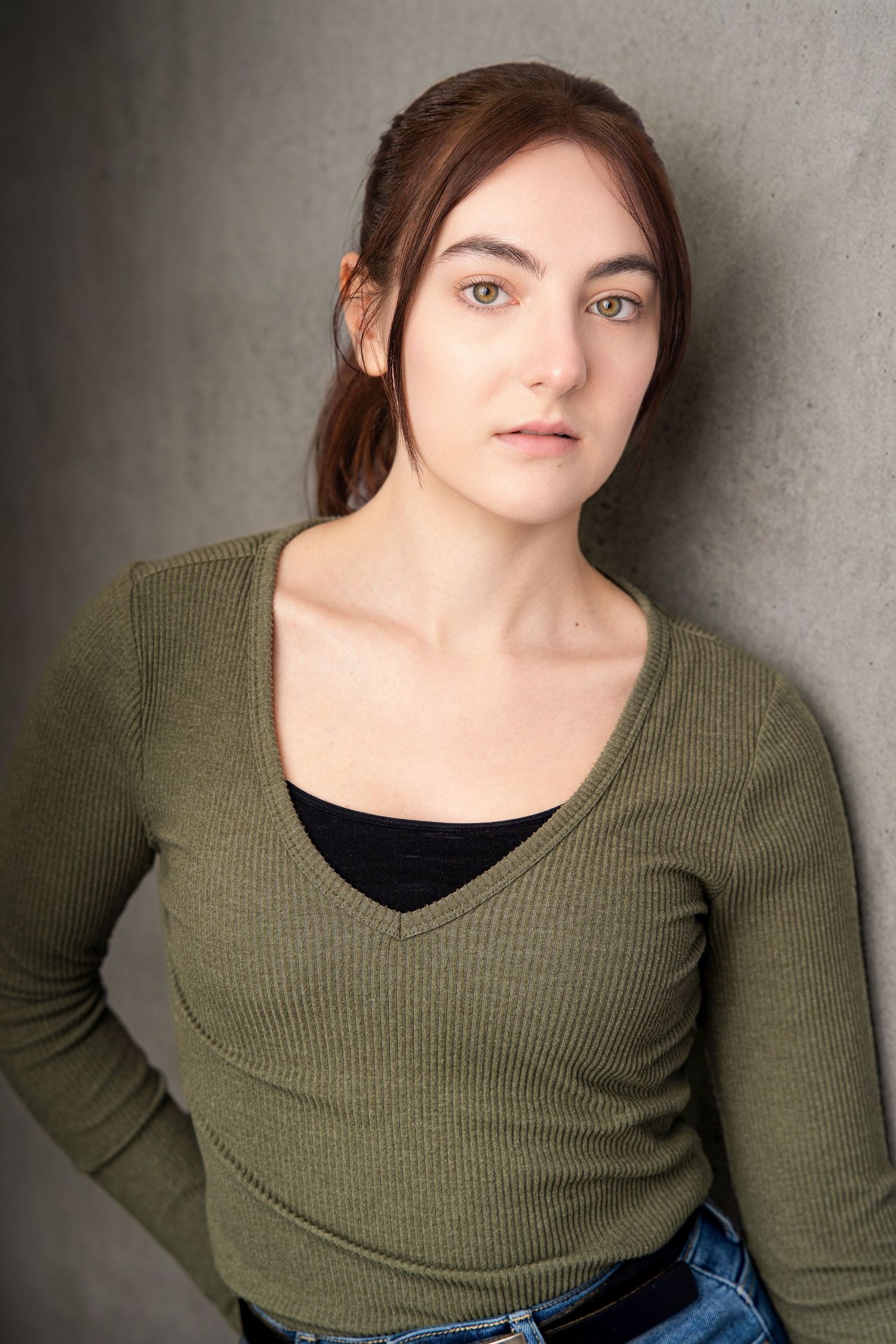 Young woman with auburn hair wearing a green V-neck sweater and jeans leaning against a gray wall.
