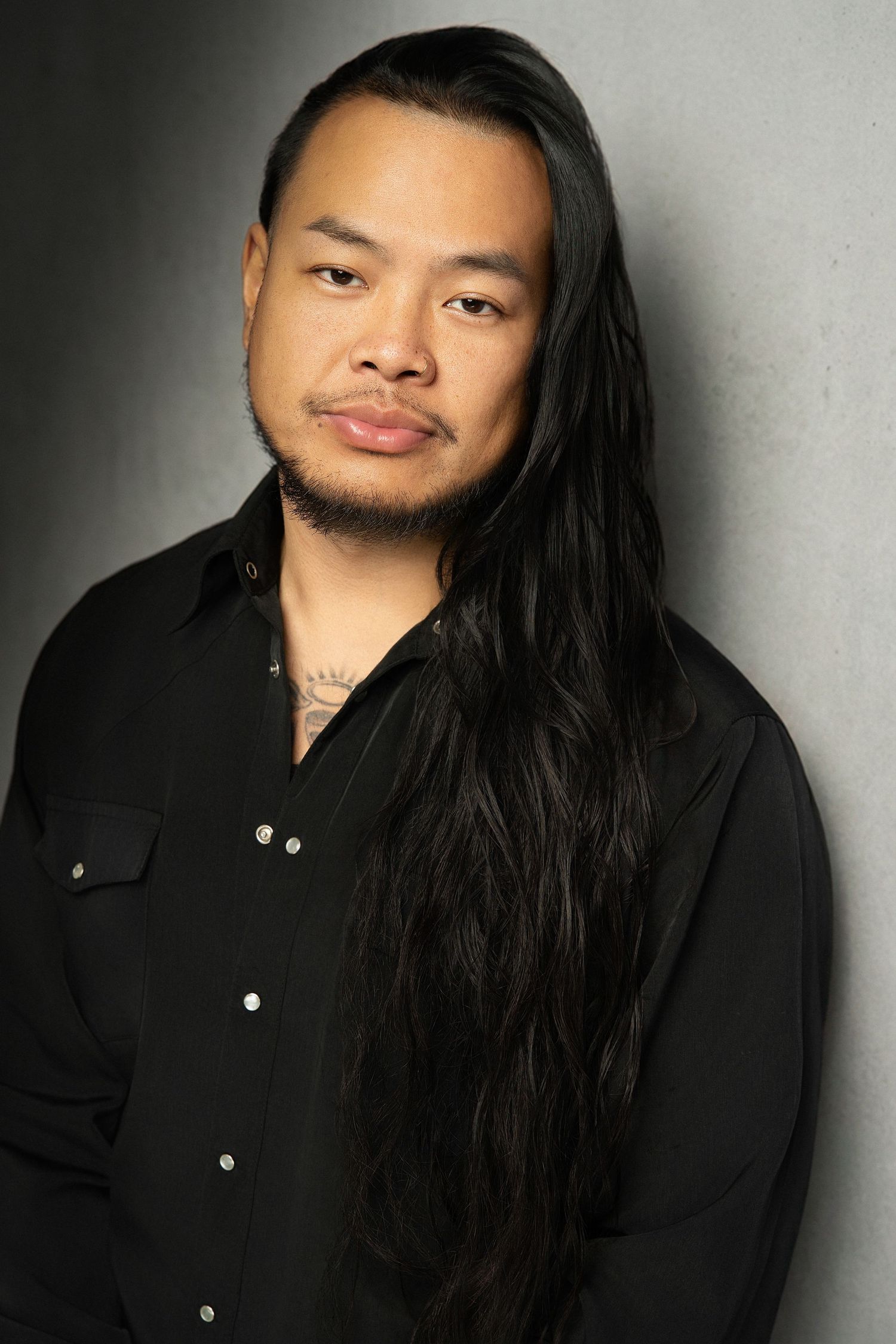 Asian man with very long black hair wearing a black shirt leaning against a gray studio backdrop.