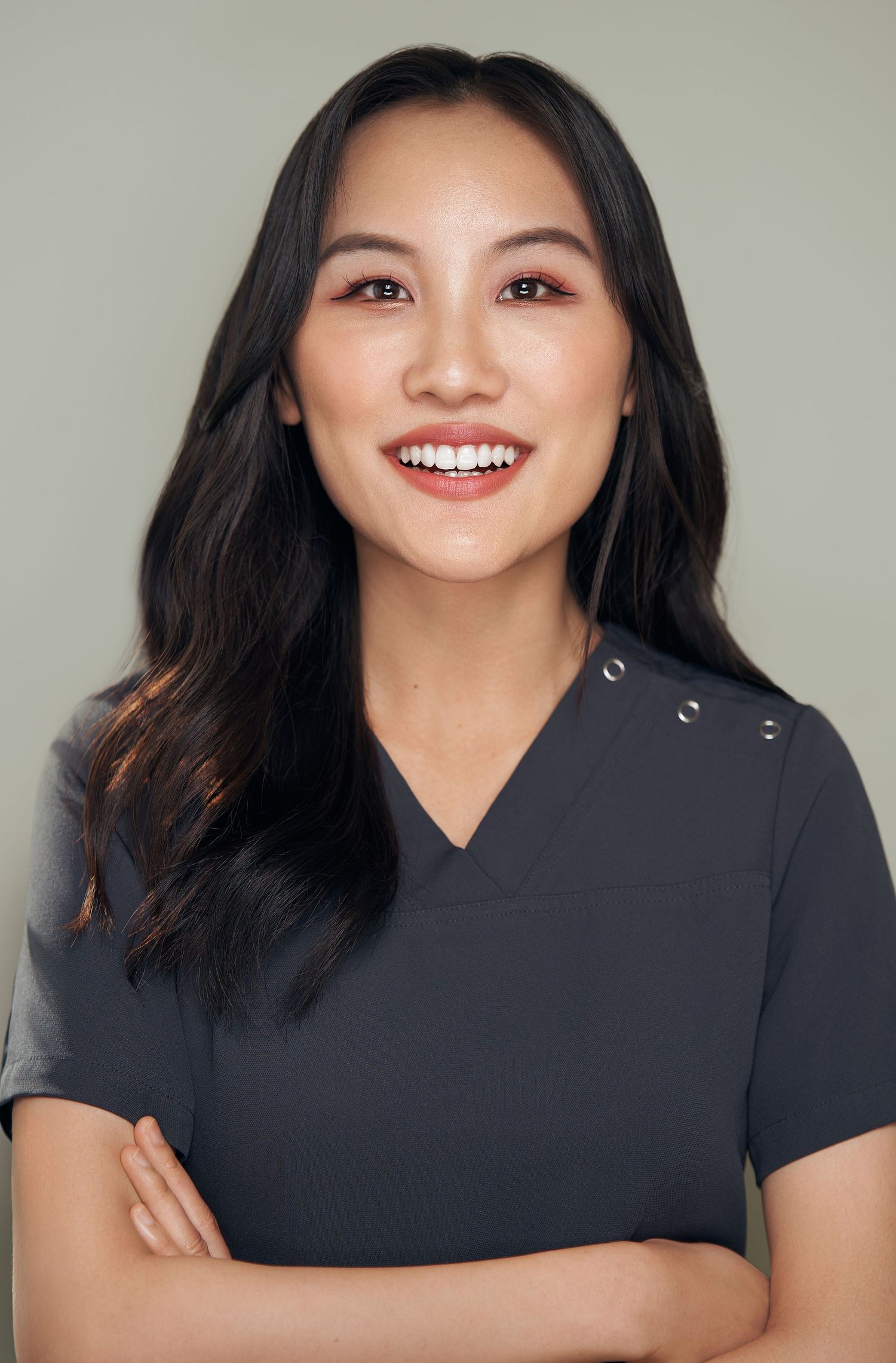 Smiling Asian woman with crossed arms wearing a dark scrub top in a professional headshot.