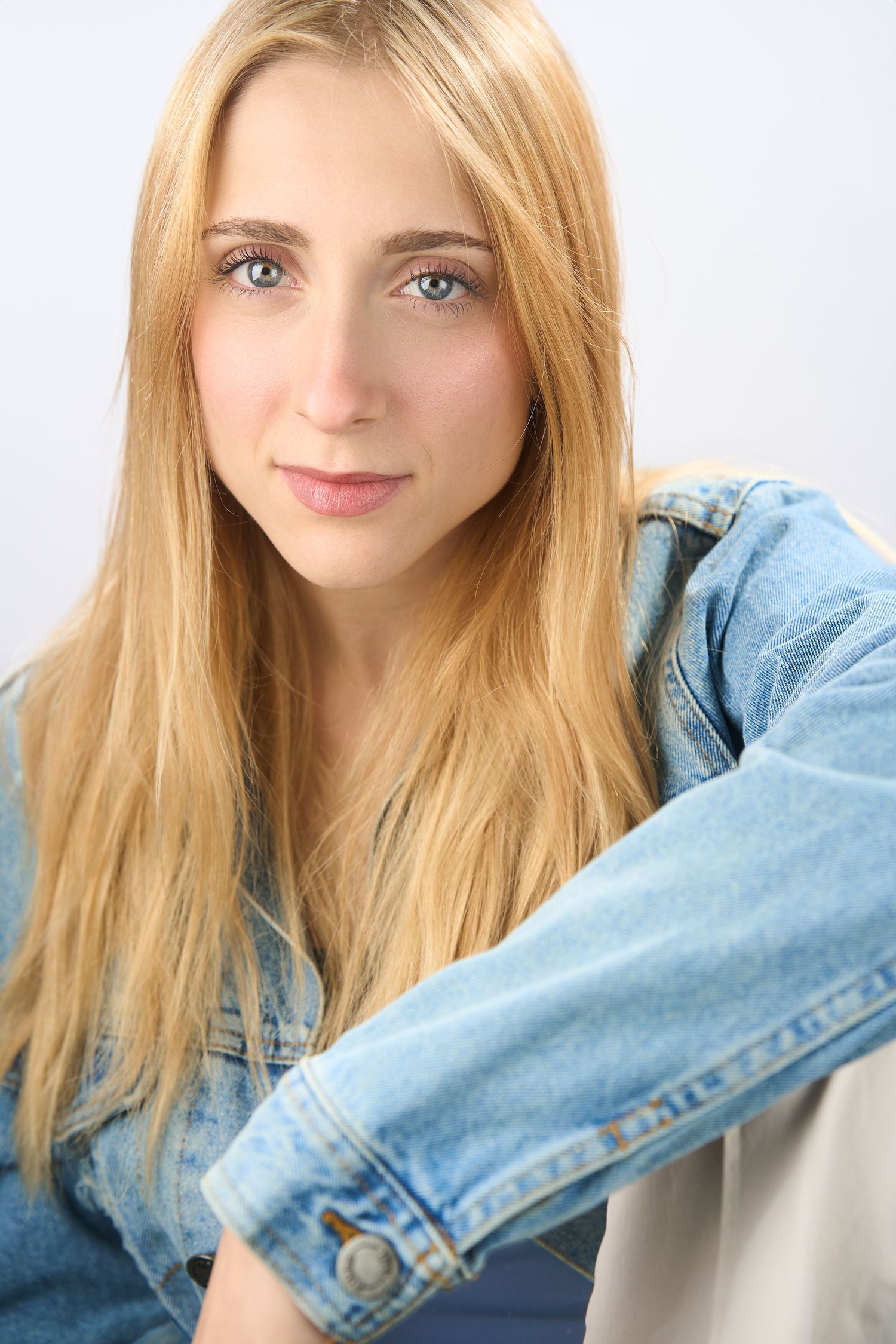 Young blonde woman with blue eyes wearing a denim jacket, posing for a portrait against a white background.