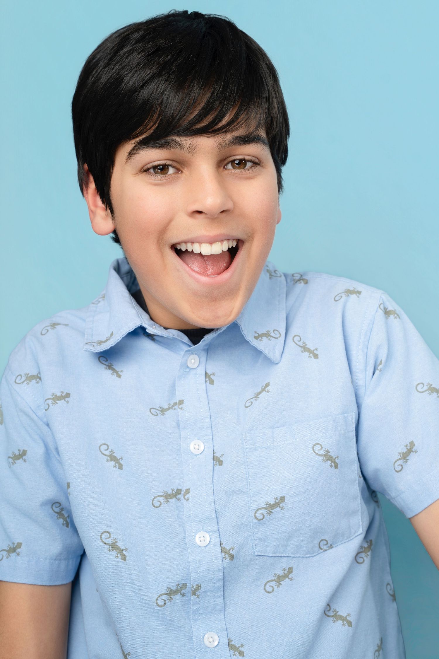 Smiling preteen boy with dark hair wearing a light blue lizard-print button-up shirt against a blue background.