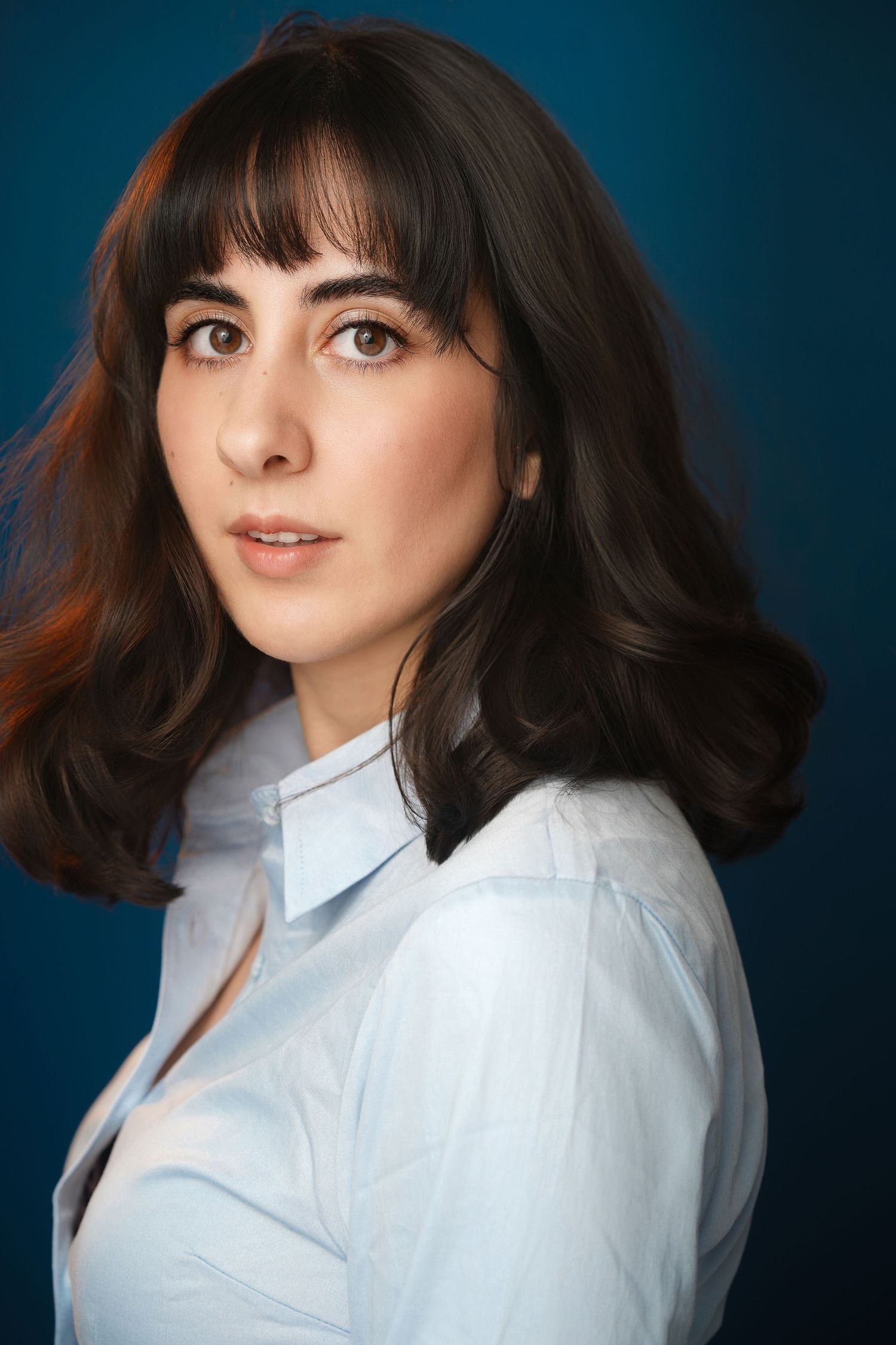 Professional headshot of a young woman with dark wavy hair and bangs wearing a light blue shirt against a blue background.