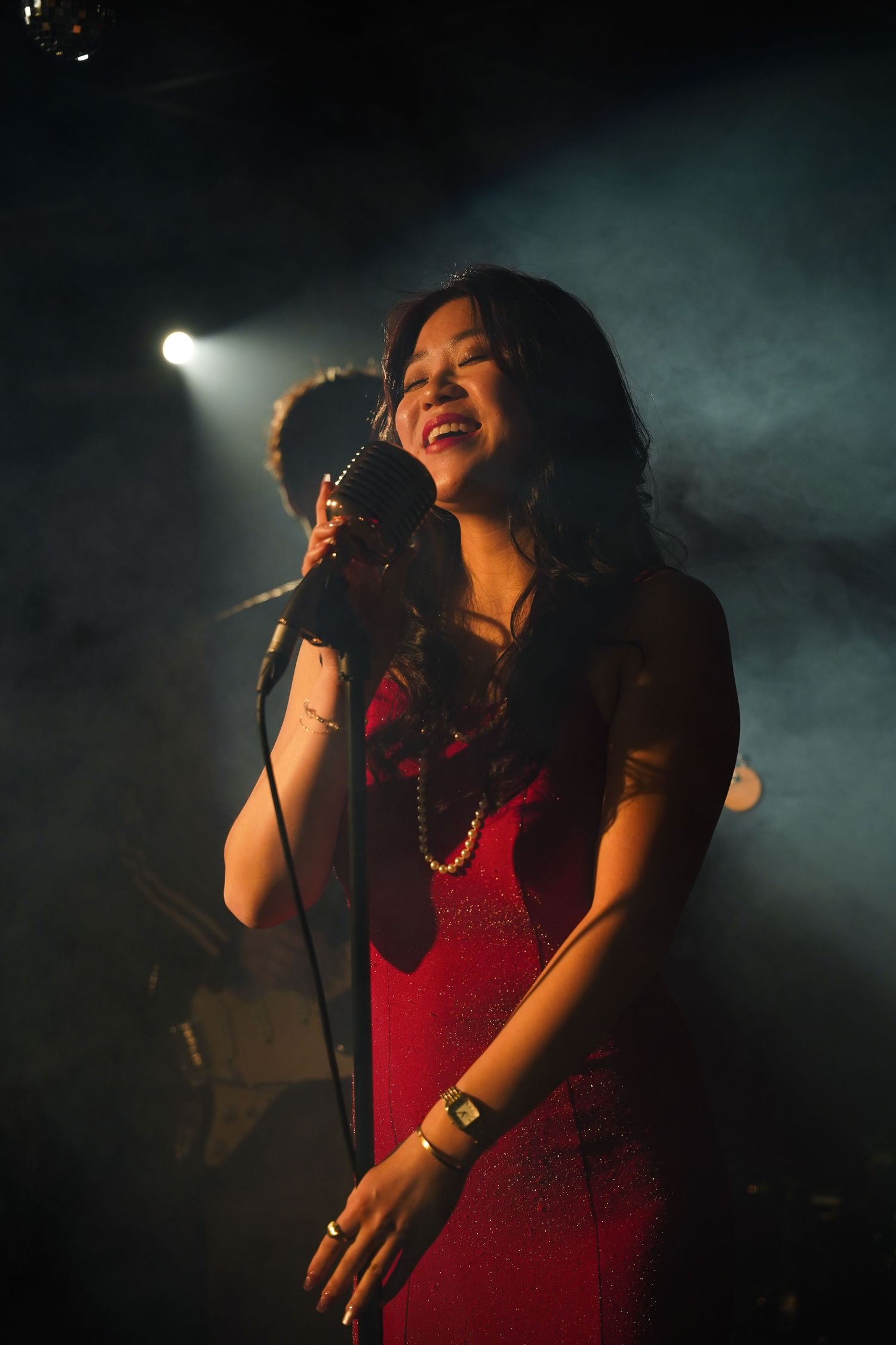 Asian woman in red dress singing passionately into vintage microphone on a smoky stage.