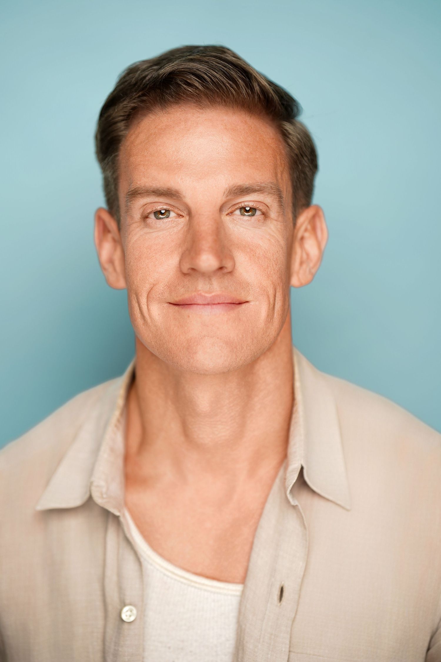 Professional headshot of a smiling middle-aged man with short brown hair wearing a beige linen shirt on blue background.