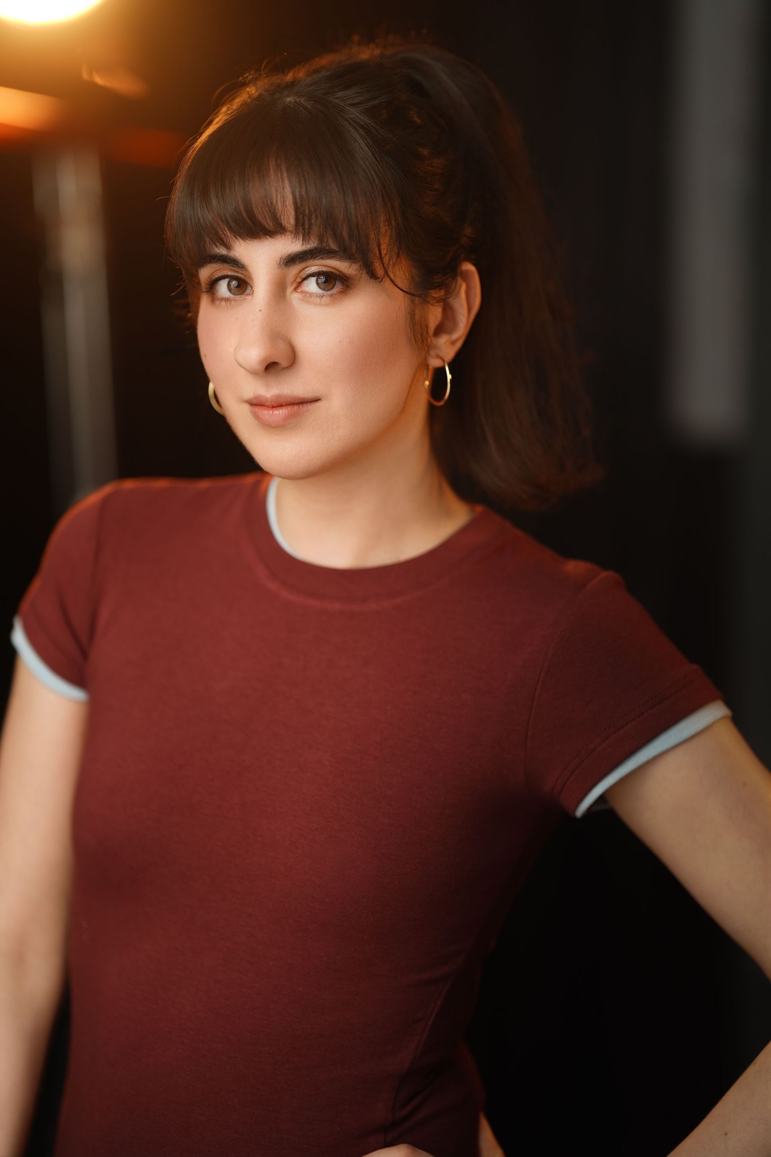 Young woman in a burgundy t-shirt with hoop earrings posing in a warmly lit studio setting.