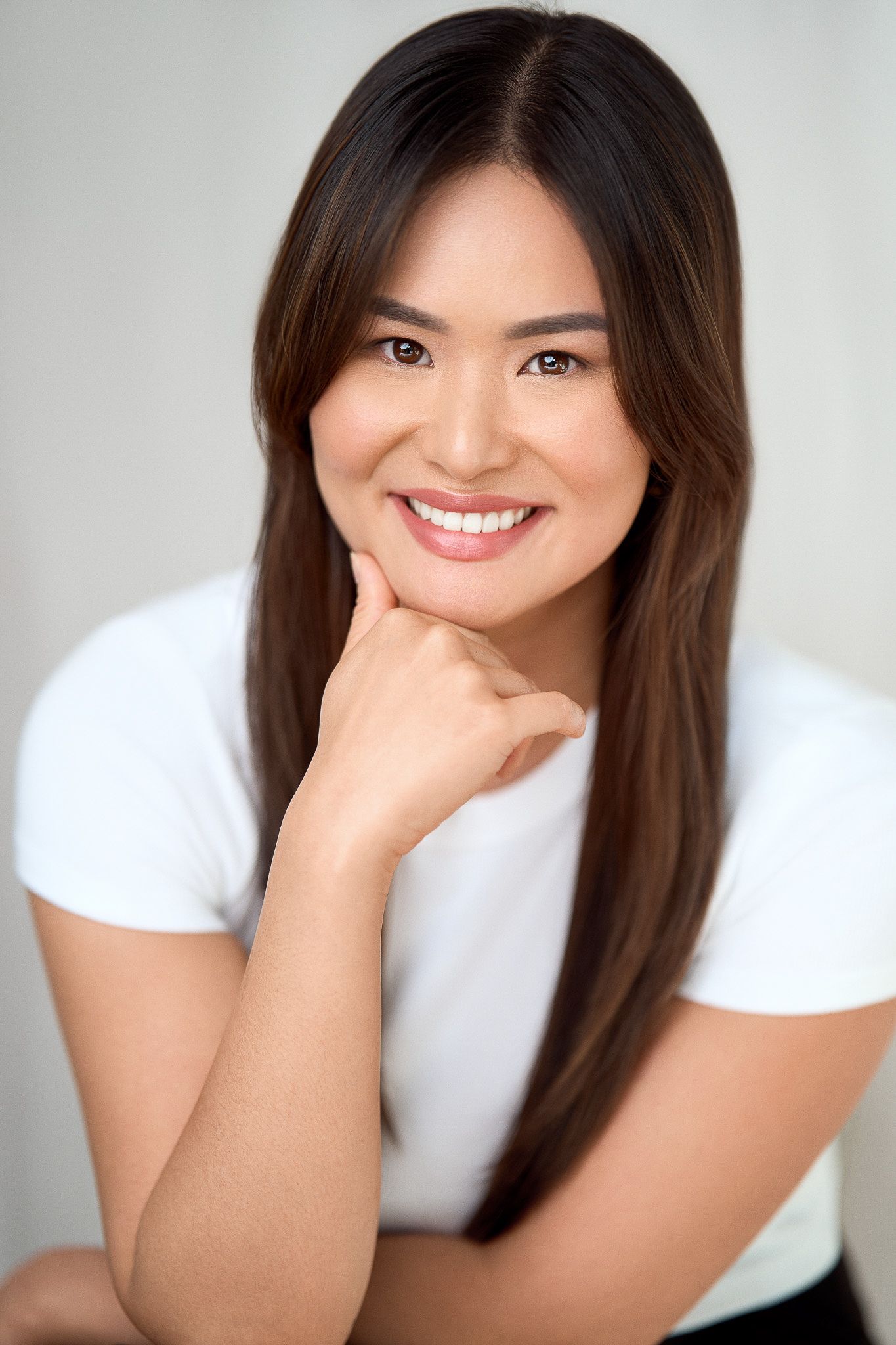 Professional headshot of a smiling Asian woman with long dark hair resting chin on hand.
