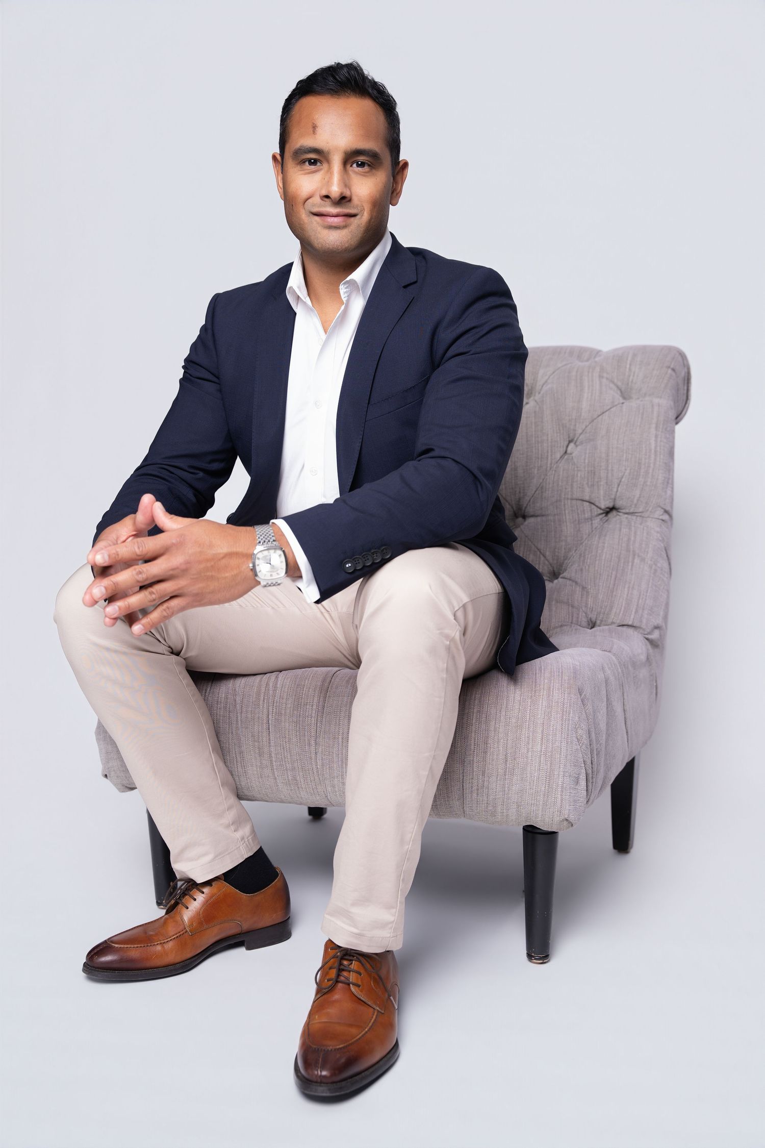 Professional businessman in navy blazer sitting confidently in a grey armchair against a neutral grey background.