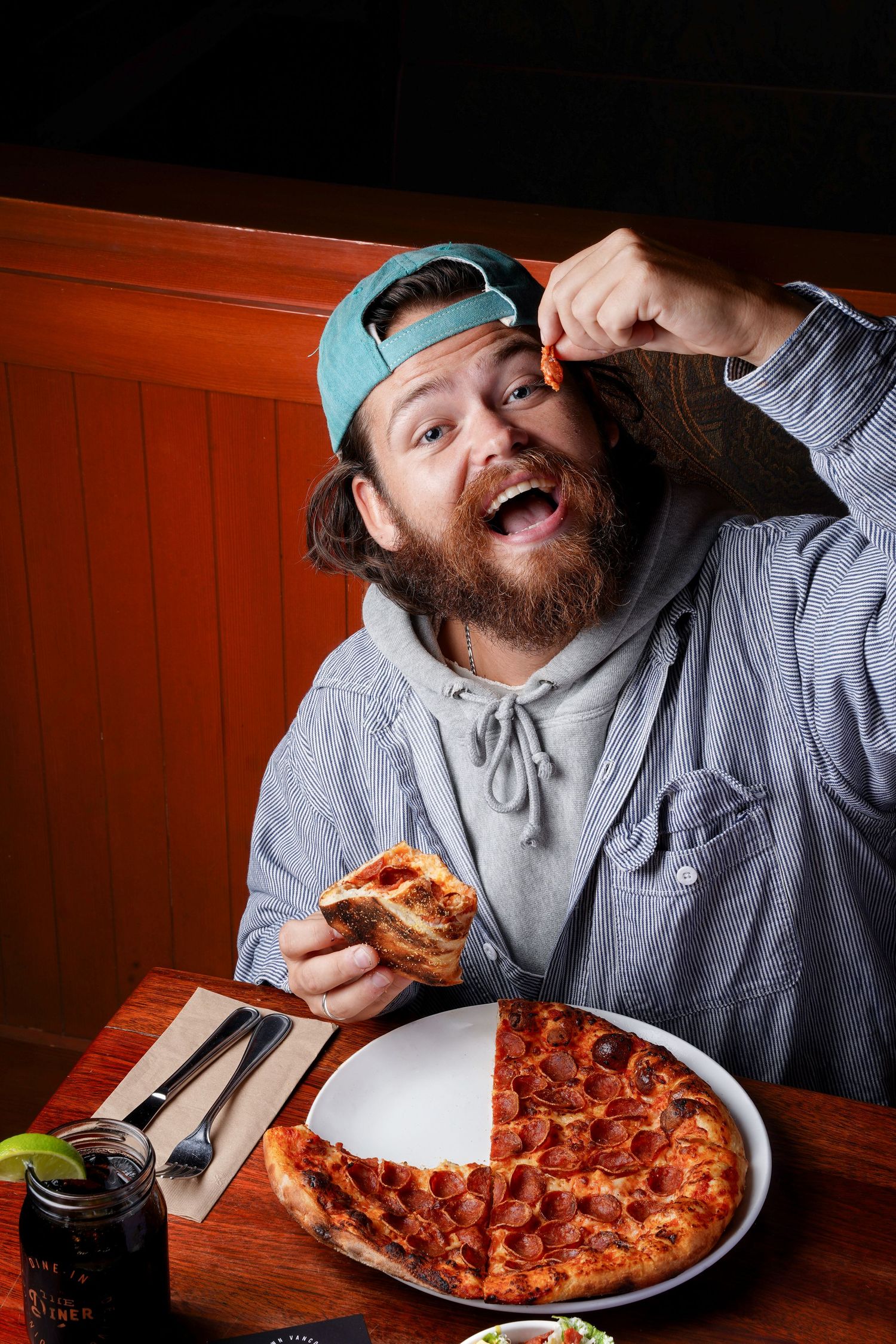 Bearded man in cap enthusiastically eating pepperoni pizza at a restaurant booth.
