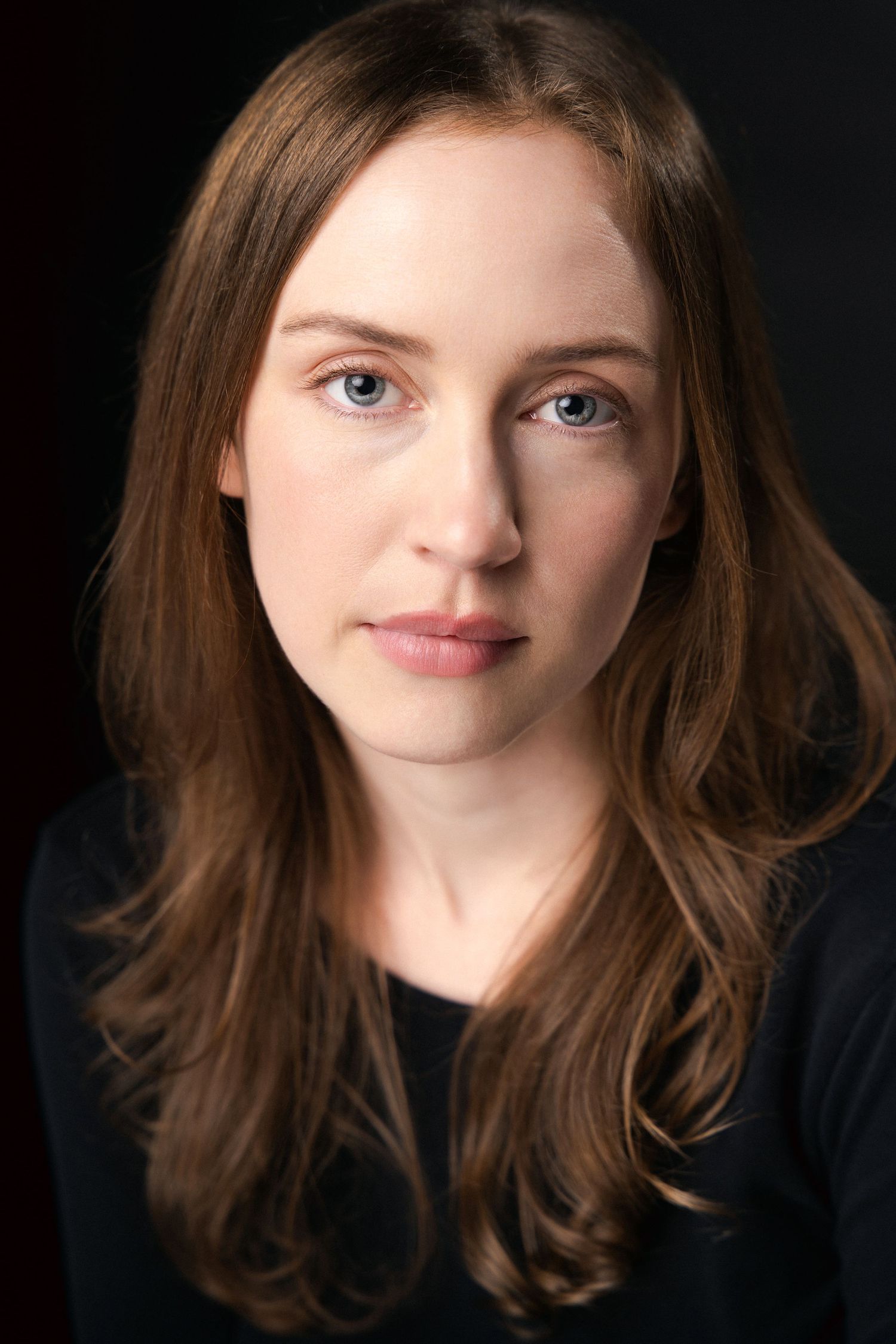 Young woman with long brown hair and blue eyes wearing a black top against a dark background.
