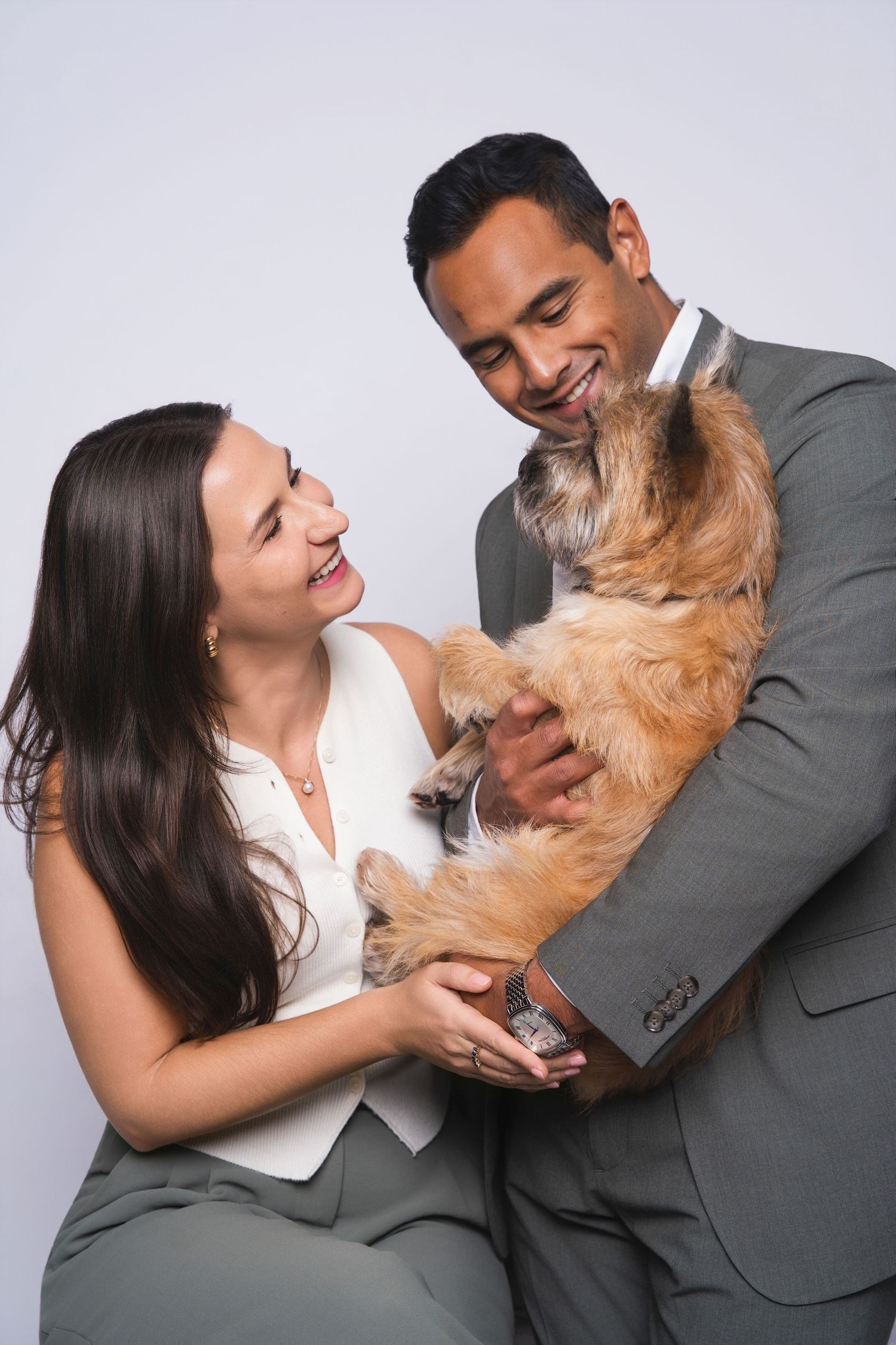 Smiling couple in formal attire holding an adorable fluffy brown dog in a studio portrait.