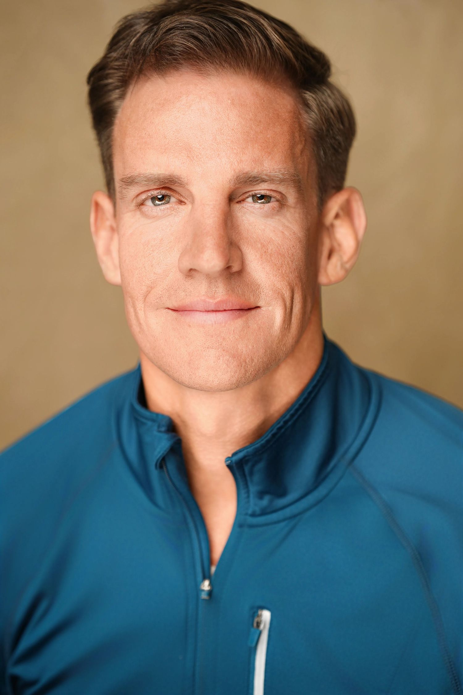 Fit middle-aged man with brown hair wearing a blue athletic zip-up jacket smiling against a tan background.