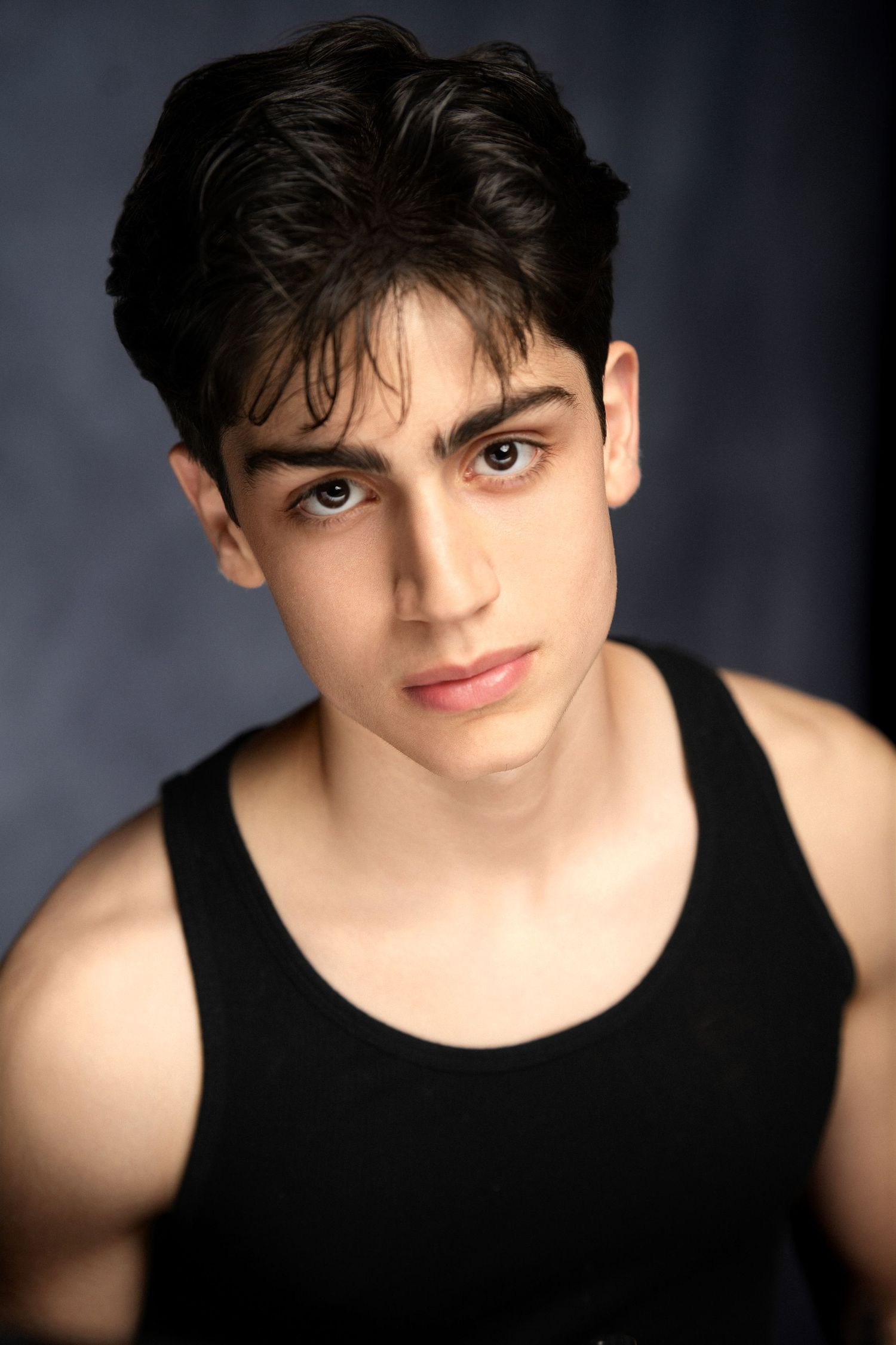 Young man with dark hair in black tank top posing against a grey studio background.