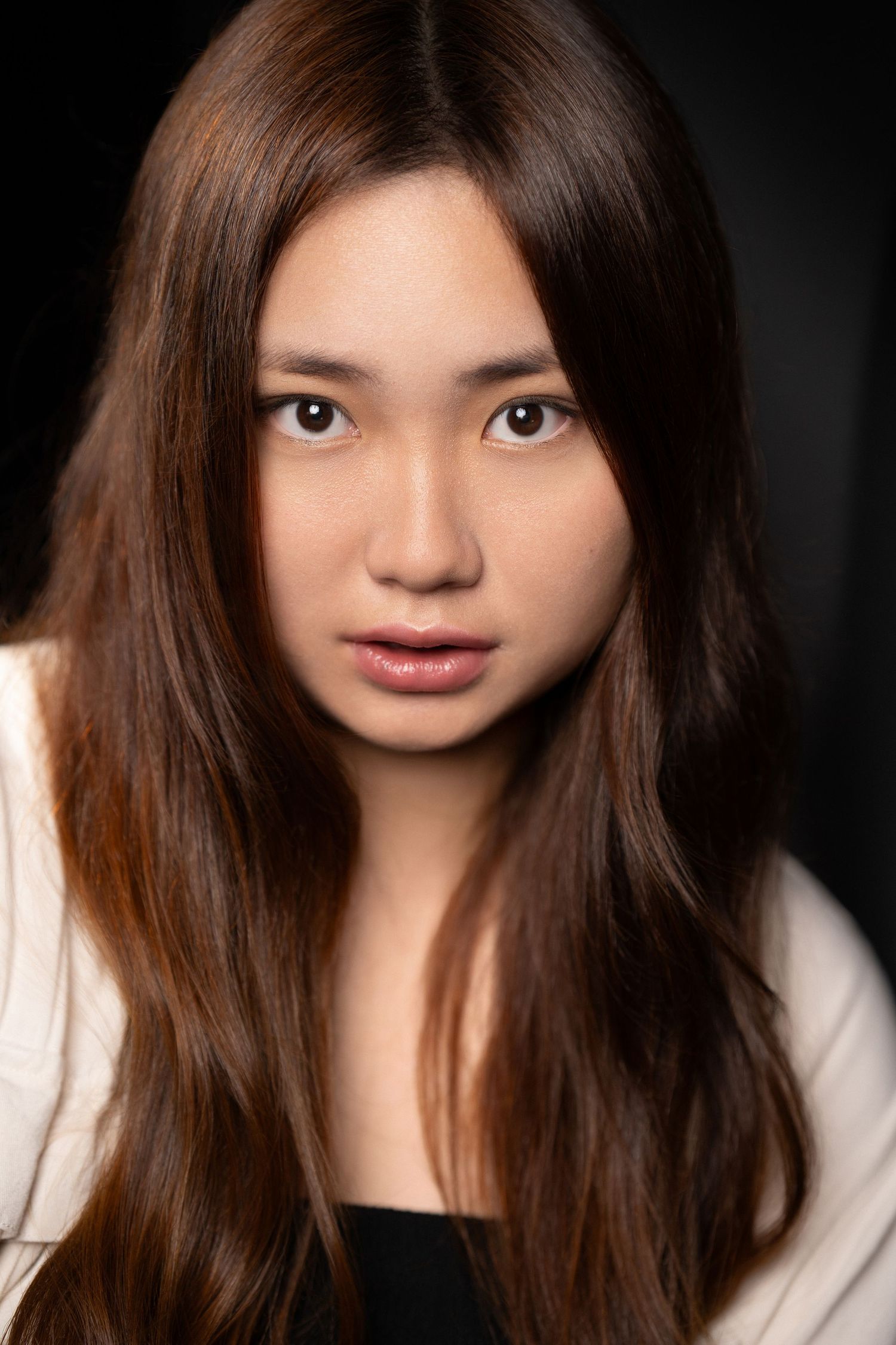 Young Asian woman with long brown hair wearing a white jacket posing against a dark black background.
