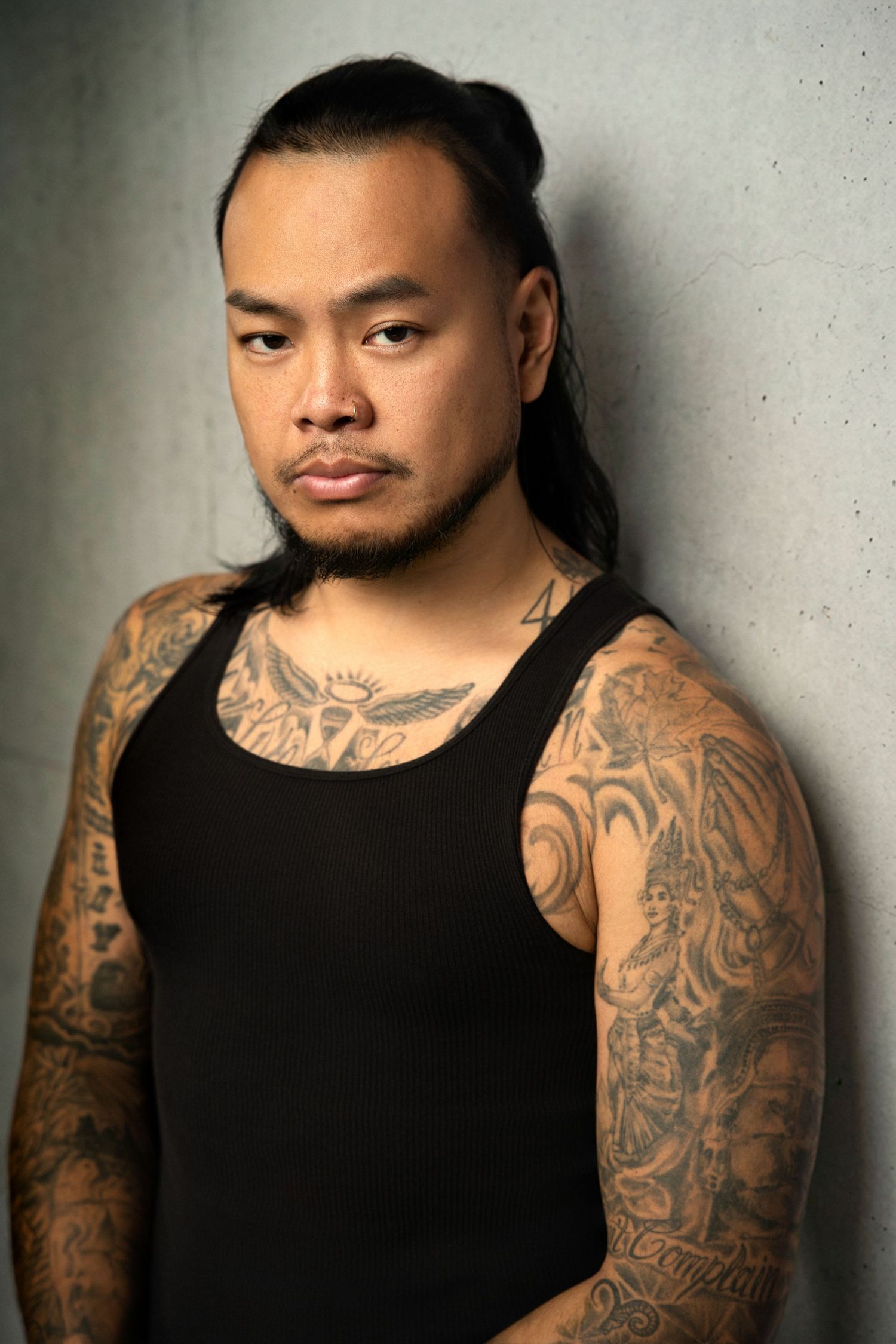 Tattooed Asian man with long dark hair in a black tank top posing against a gray concrete wall.