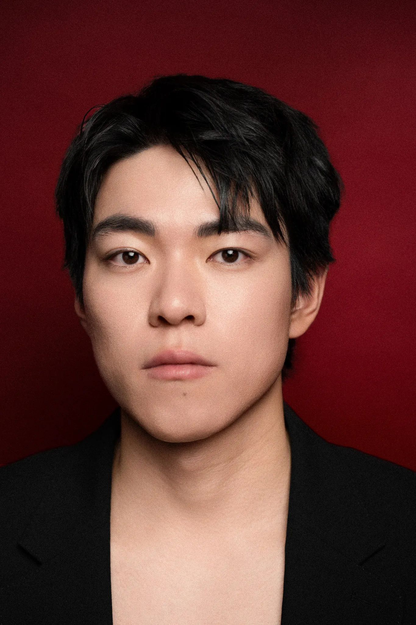 Young Asian man with dark hair wearing a black jacket against a deep red background.