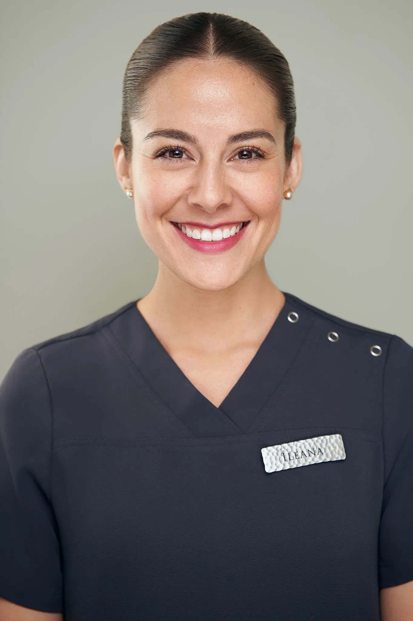Smiling female healthcare professional in dark scrubs with name badge on neutral background.