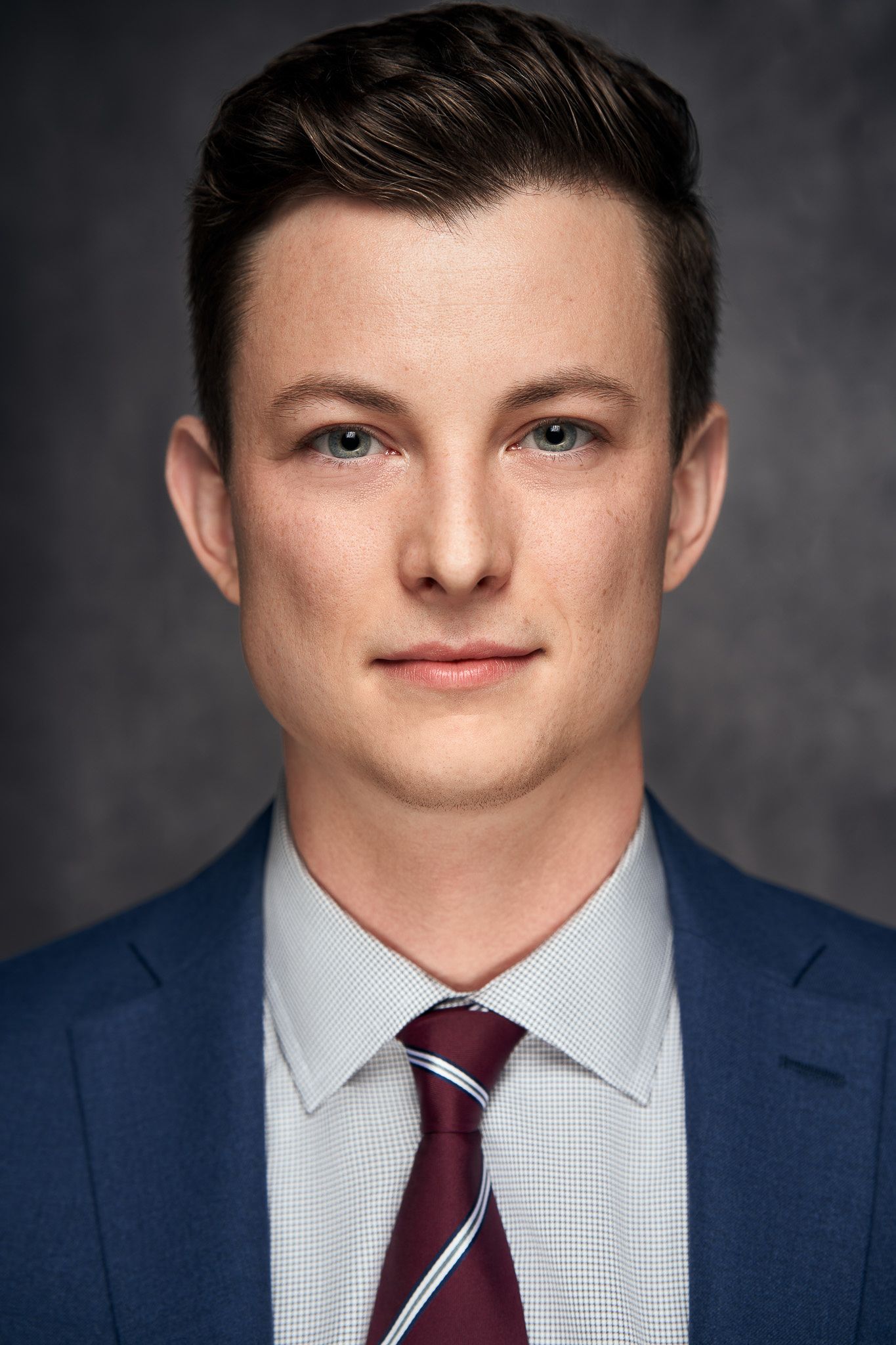 Professional headshot of a young man in a blue suit and burgundy tie against a dark background.