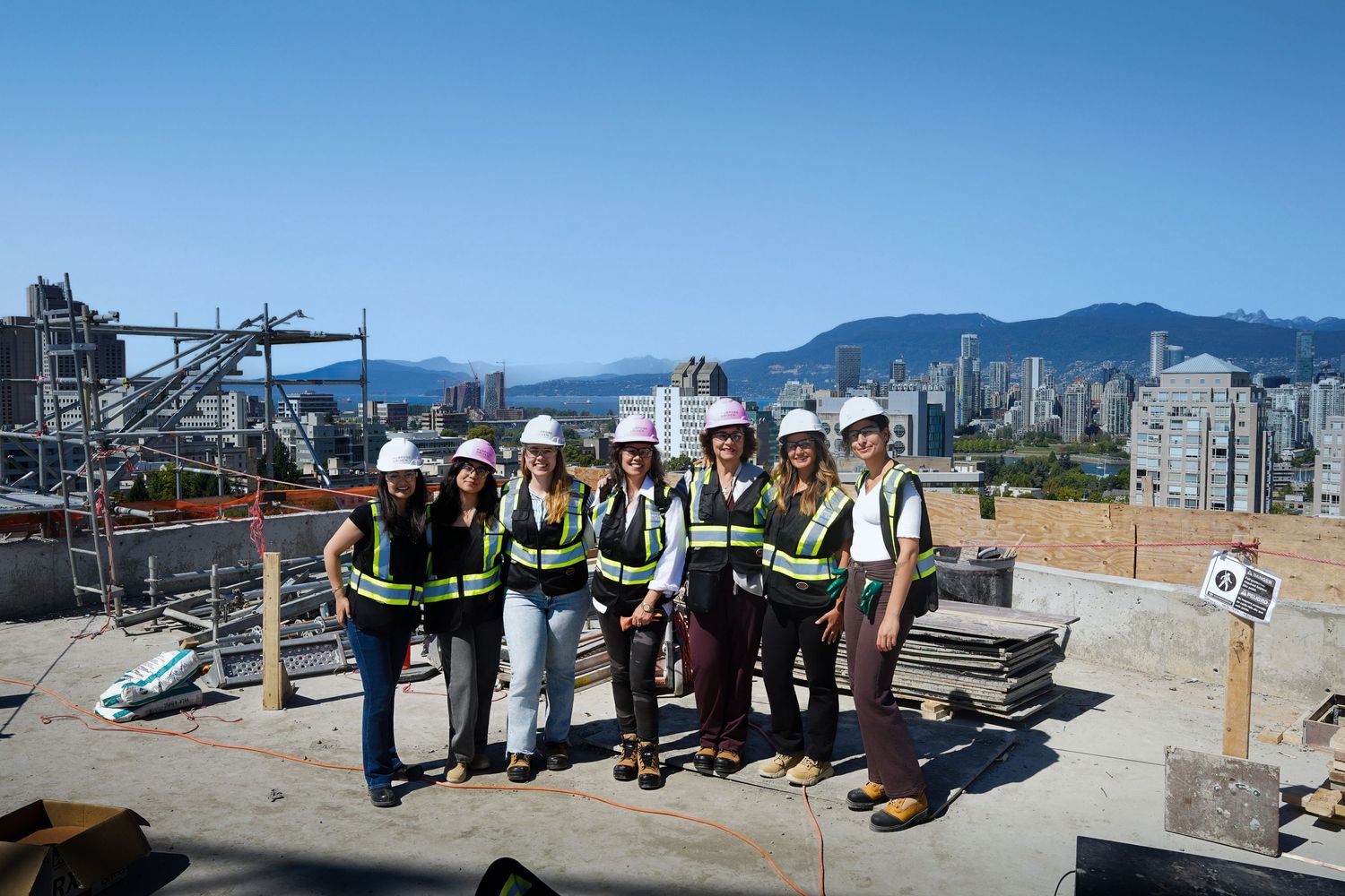 Group of seven women wearing safety vests and hard hats posing on a rooftop construction site with city skyline view.
