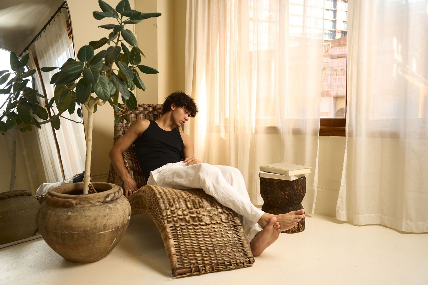 Man relaxing on wicker chaise lounge chair in bright boho living room with linen curtains and potted fiddle leaf fig.