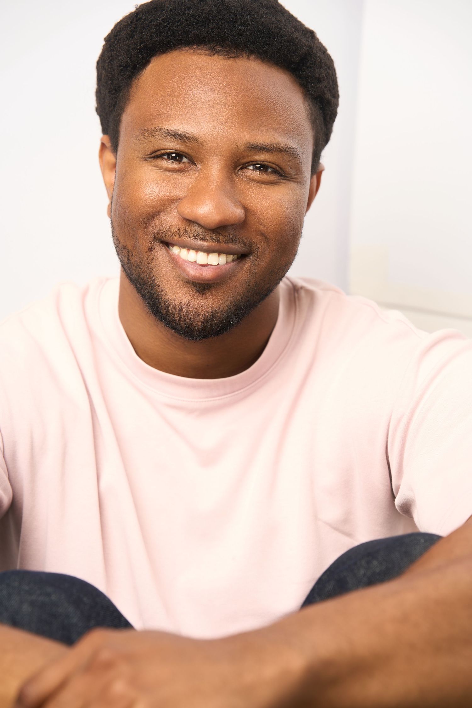 Smiling young Black man with short hair and beard wearing a light pink t-shirt, seated casually against a white background.