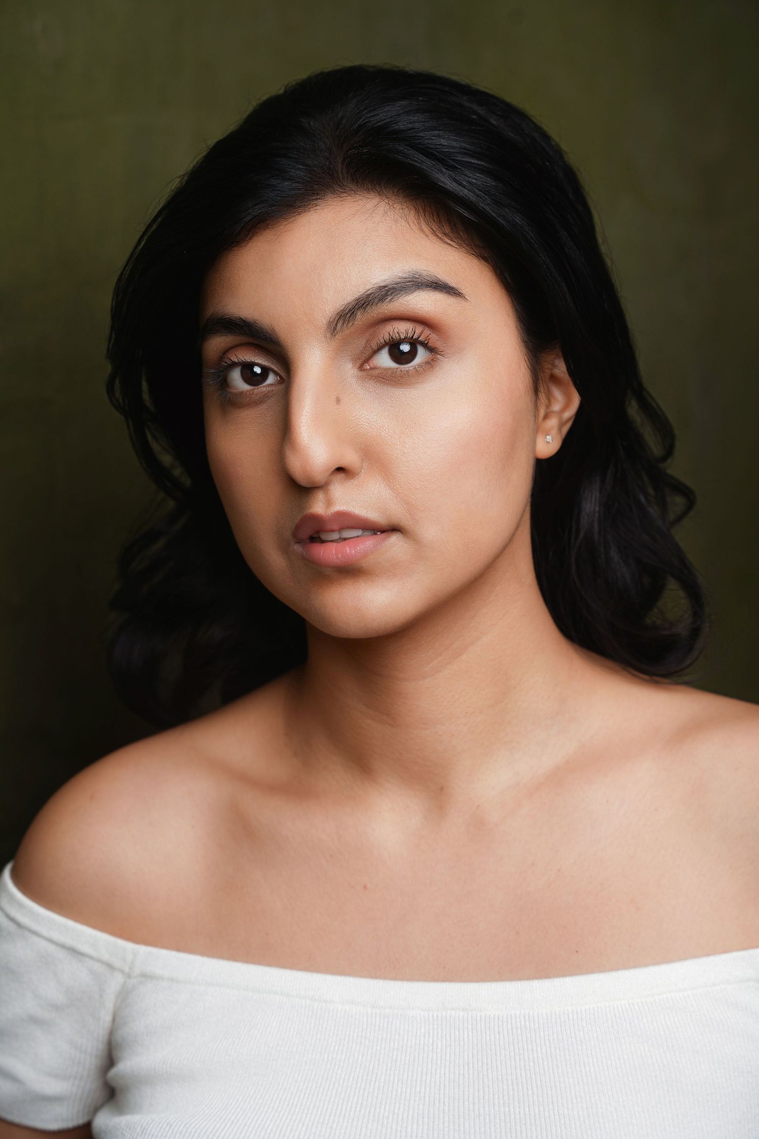 Young South Asian woman with black wavy hair wearing a white off-shoulder top against a dark green background.