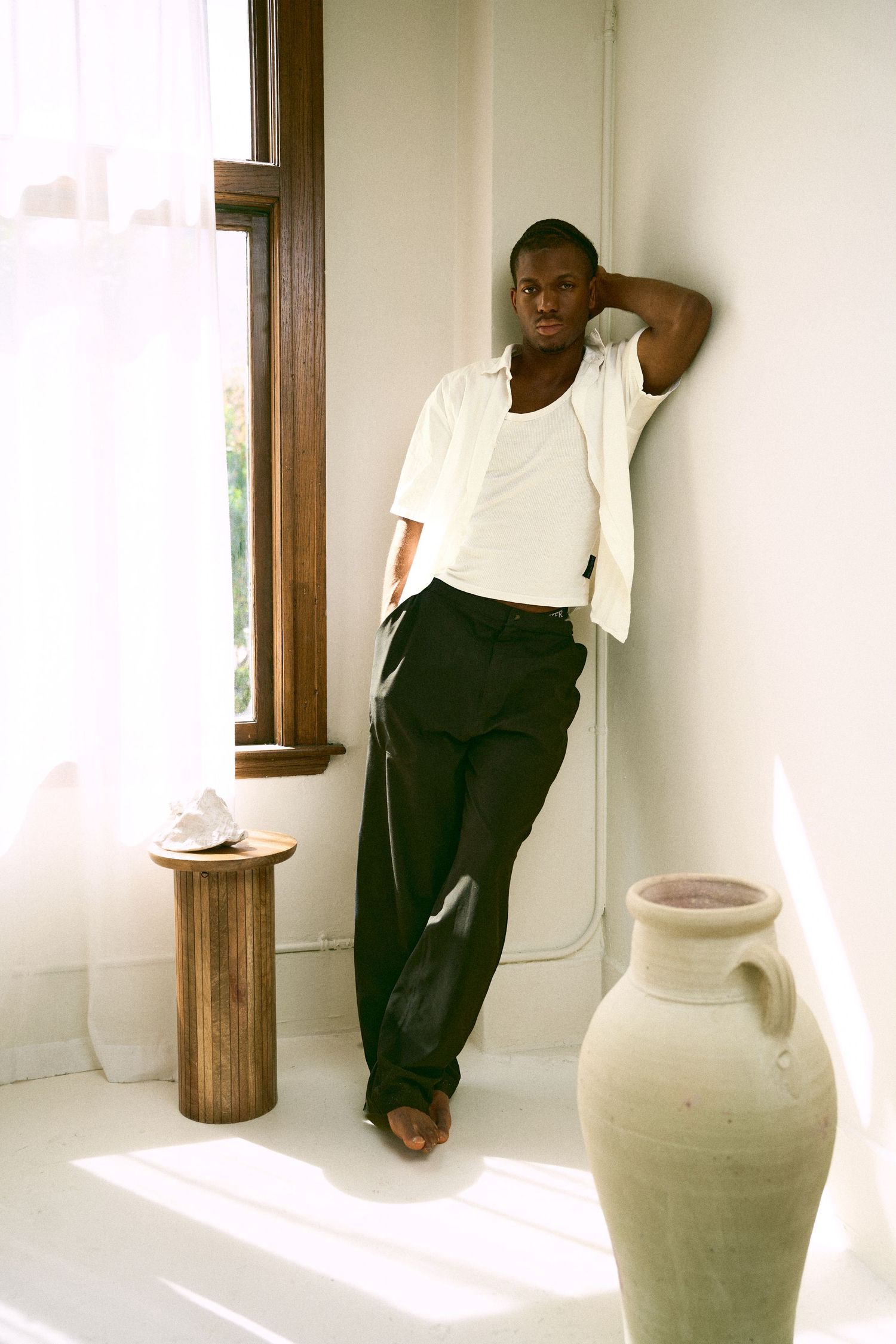 A young Black man in white shirt and black trousers leans against a white wall near a sunlit window with a ceramic vase nearby.