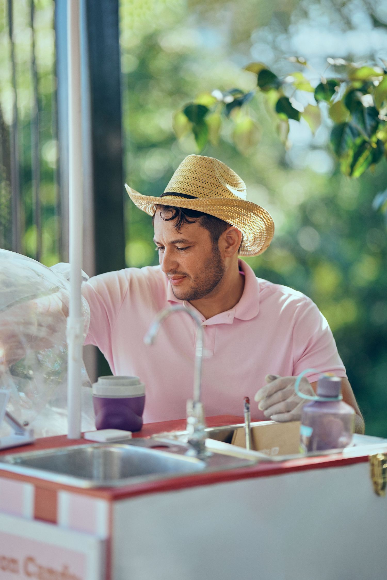 Man in pink polo shirt and straw hat working at an outdoor cotton candy vendor cart.