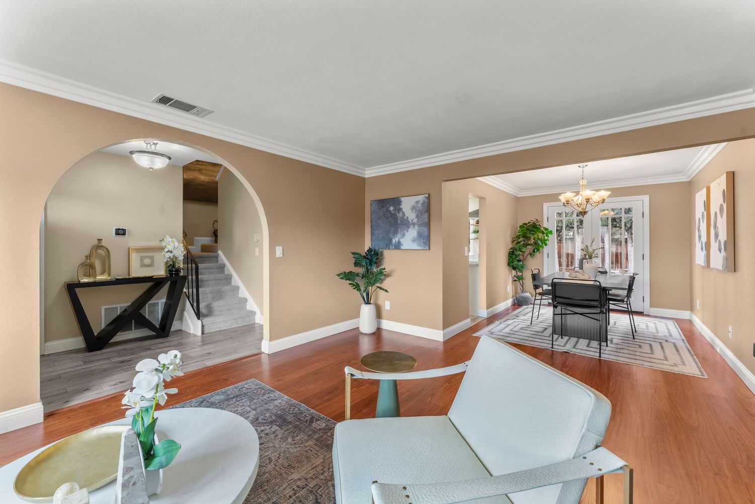 Spacious living room with arched doorway, hardwood floors, crown molding, chandelier dining area, and staircase in background.