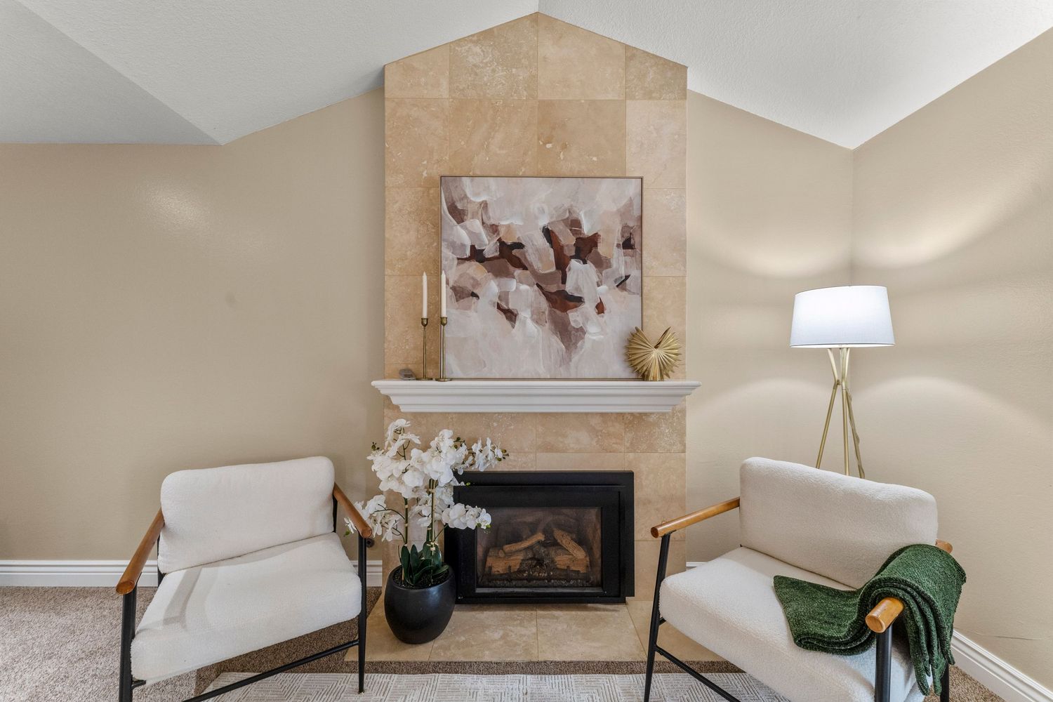 Cozy sitting area with tile-surround fireplace, vaulted ceiling, two white accent chairs, and neutral carpet flooring.