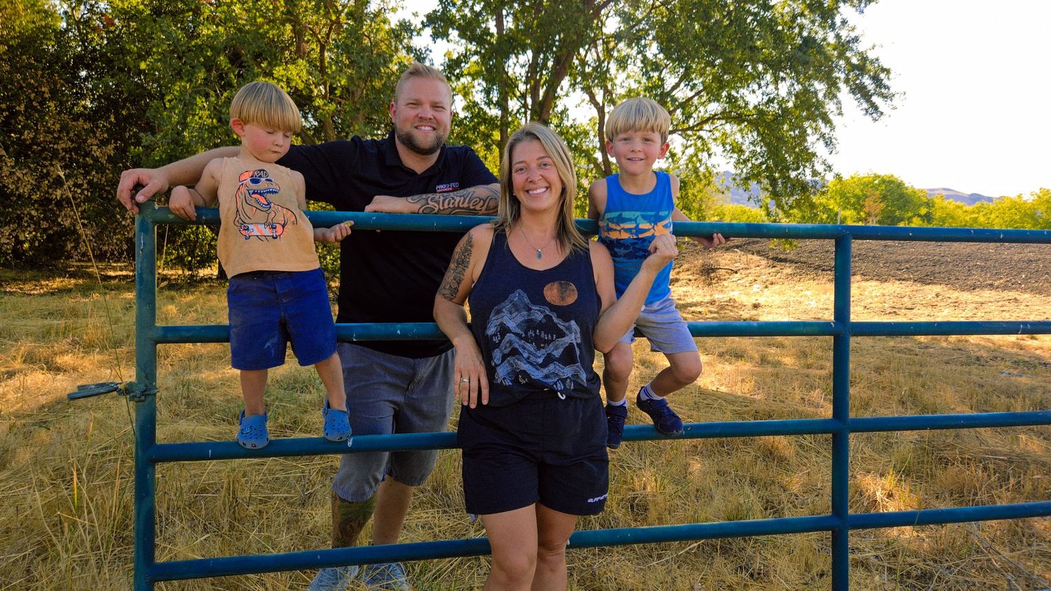 Family of four posing on a blue metal farm gate in a rural field with trees in the background.