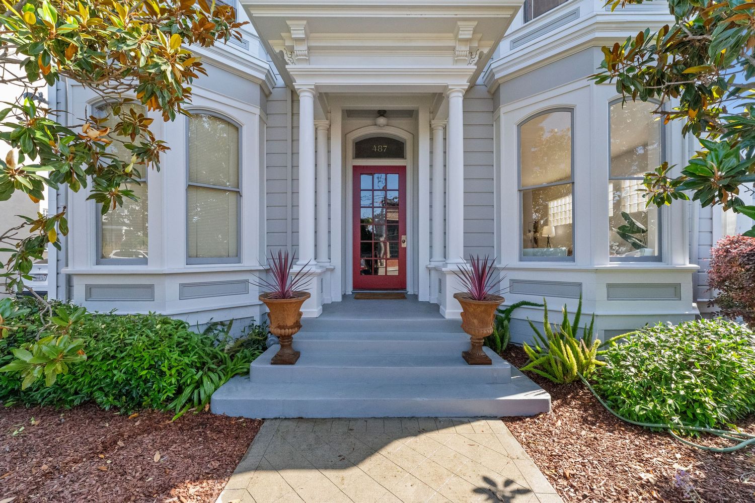 Victorian home entrance with gray exterior, red door, white columns, and lush landscaped front steps.