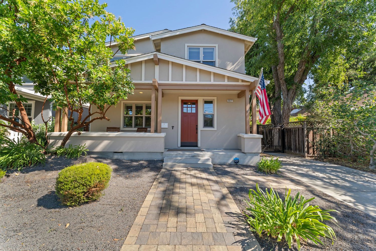 Craftsman-style home with red door, mature trees, American flag, and paver driveway.
