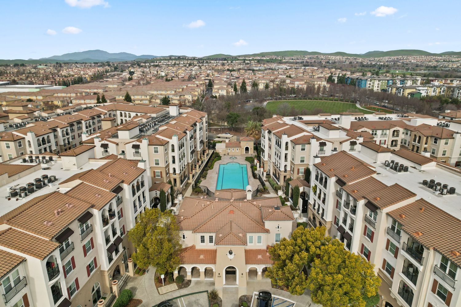 Aerial view of large Mediterranean-style apartment complex with central courtyard, pool, and mountain backdrop.