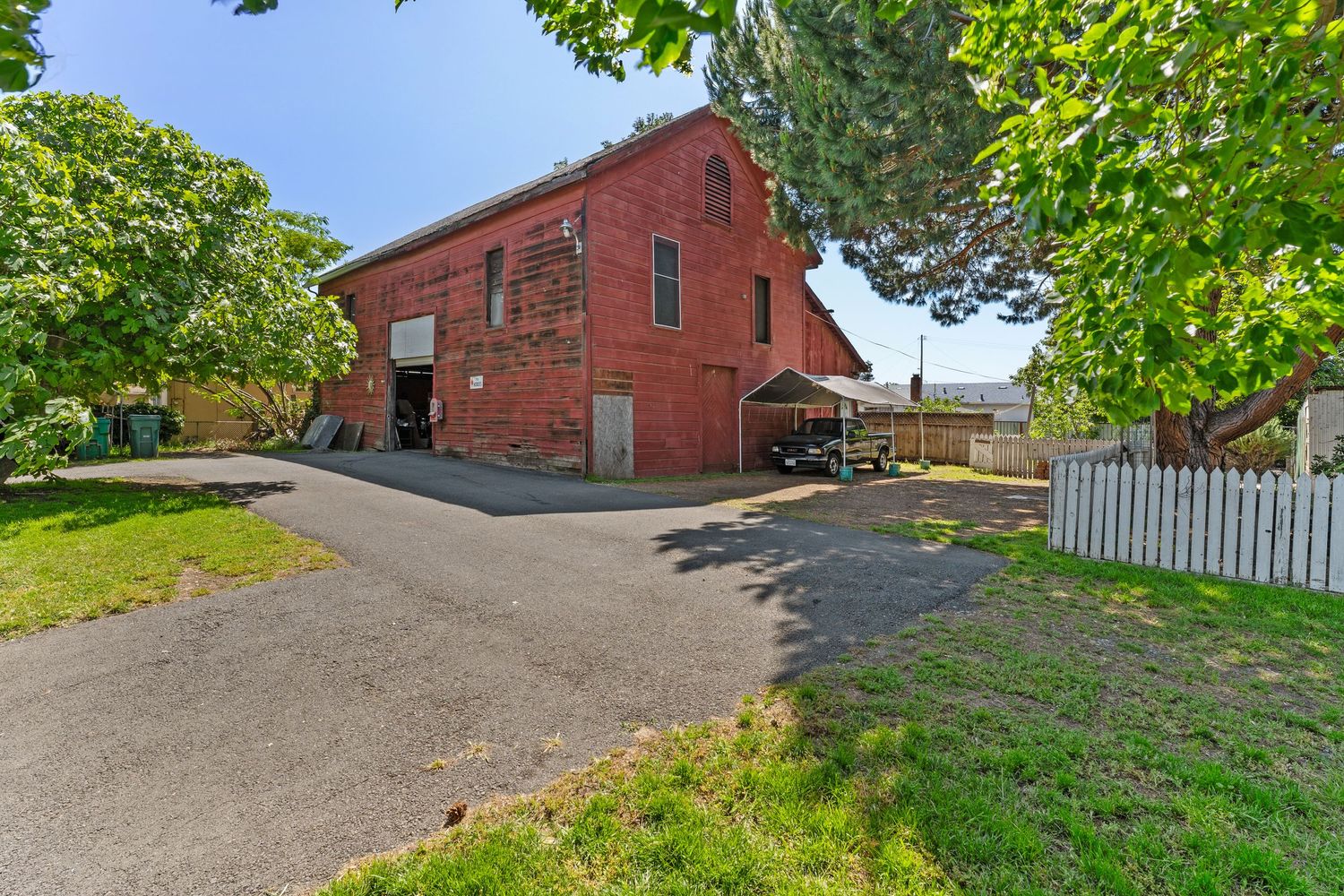 Historic red brick building with gabled roof and paved driveway, surrounded by mature trees and white picket fence.
